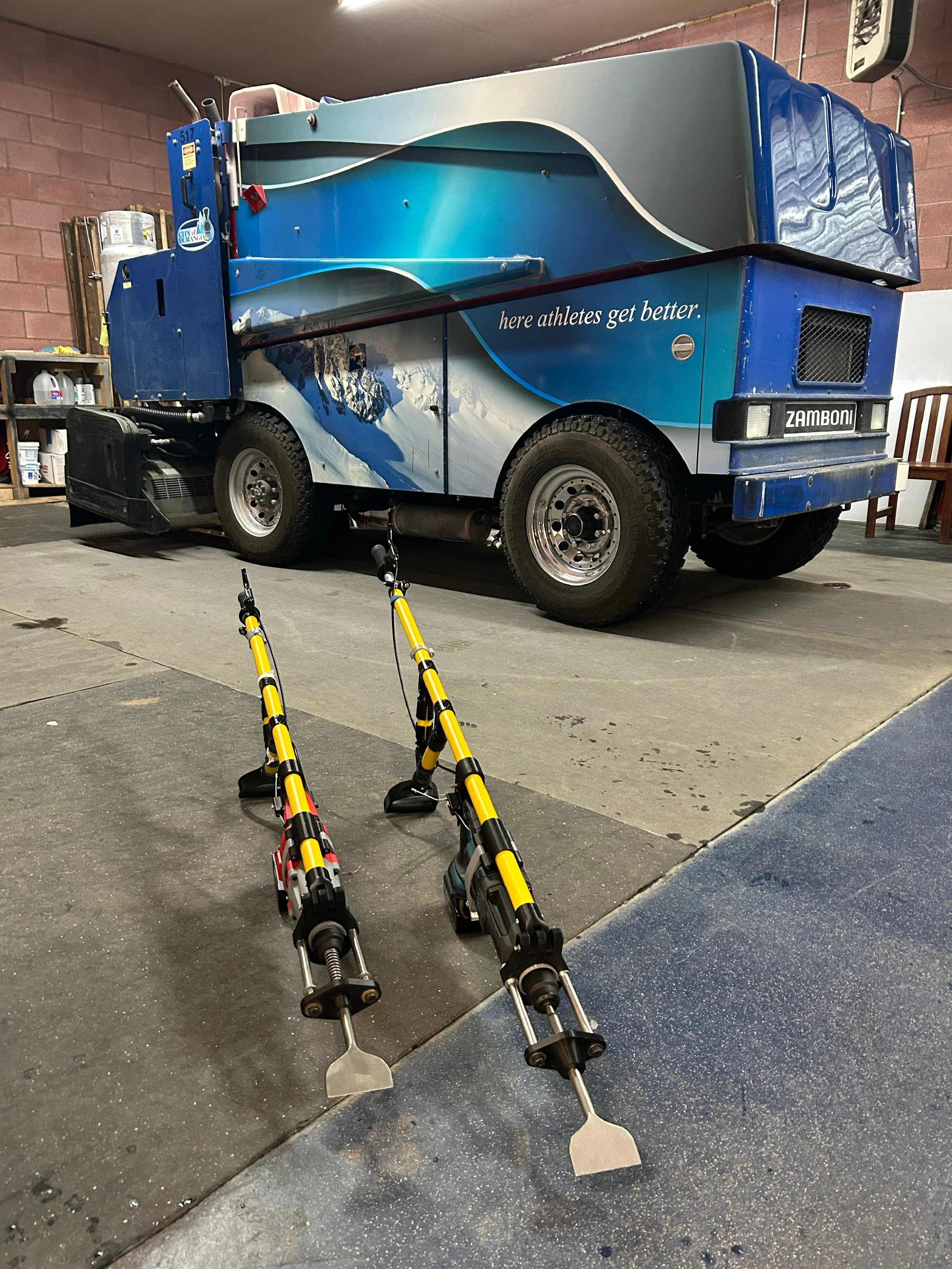 A zamboni machine parked indoors with ice chipping tools that are rotarty hammer drills placed on the floor in front of it.