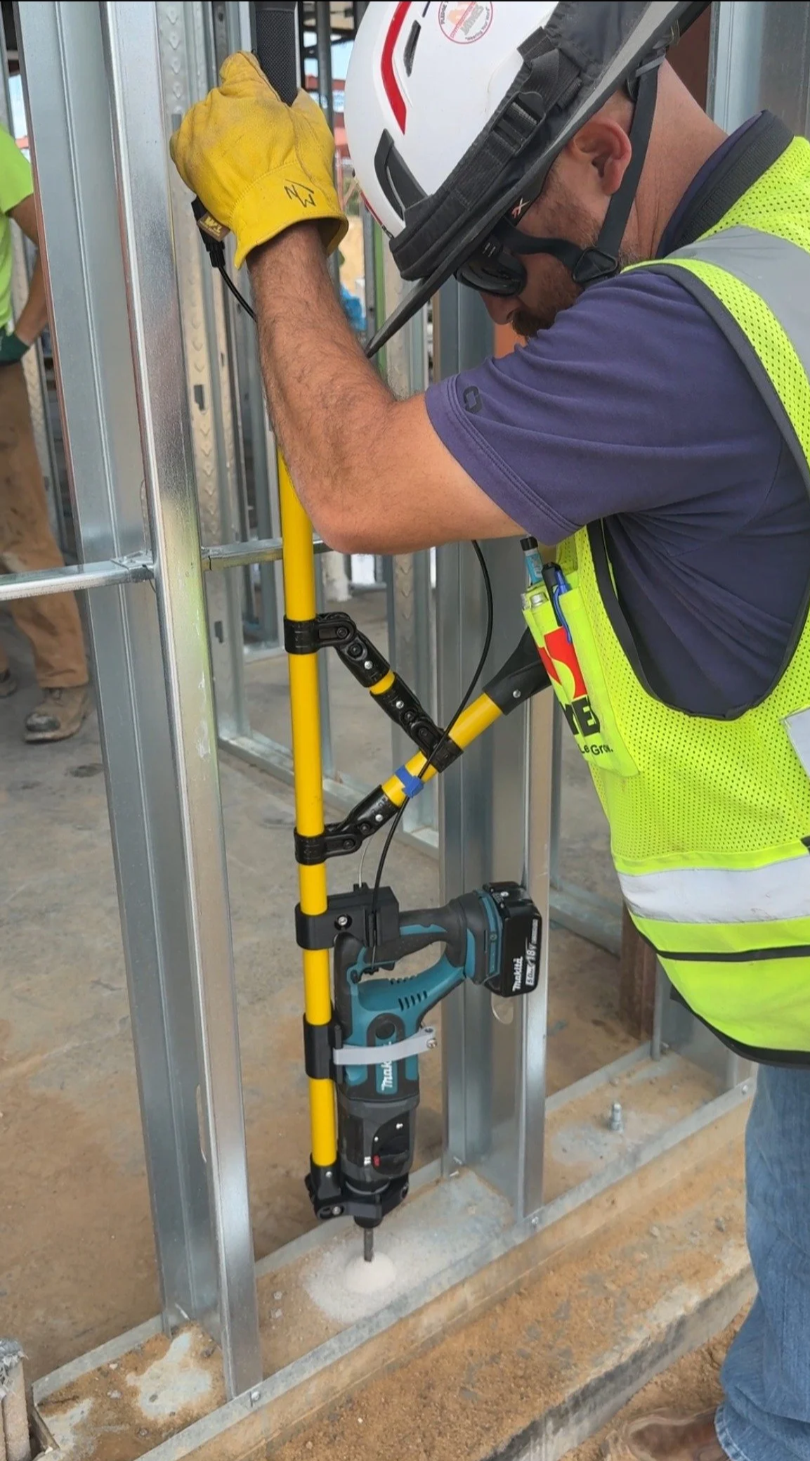 A construction worker using a cordless hammer drill to drill into a stem wall in a building under construction. The worker is wearing a yellow safety vest, a white helmet, goggles, and yellow gloves.