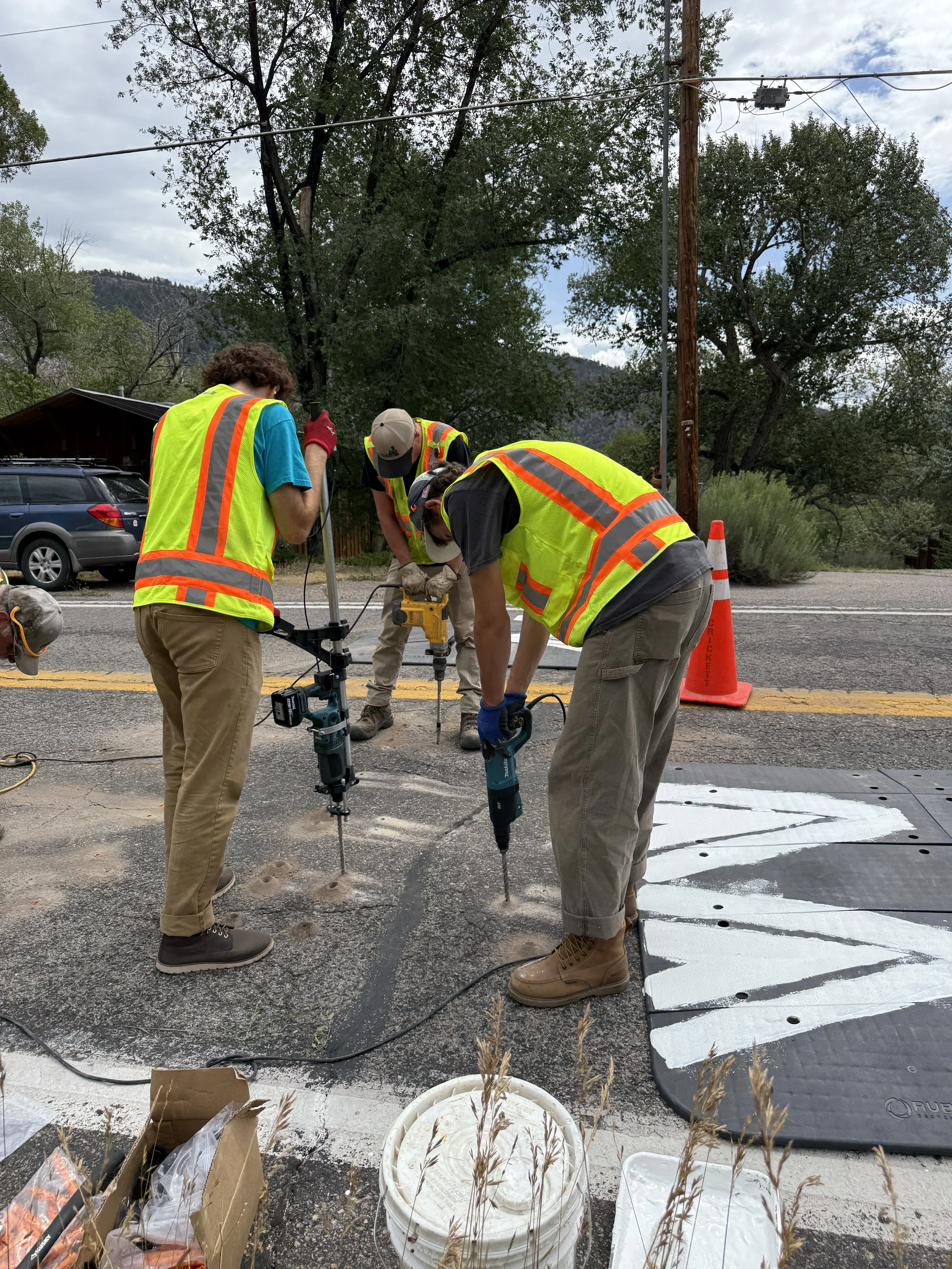Three workers in high-visibility vests and work boots are using jackhammers to break up pavement on a road.