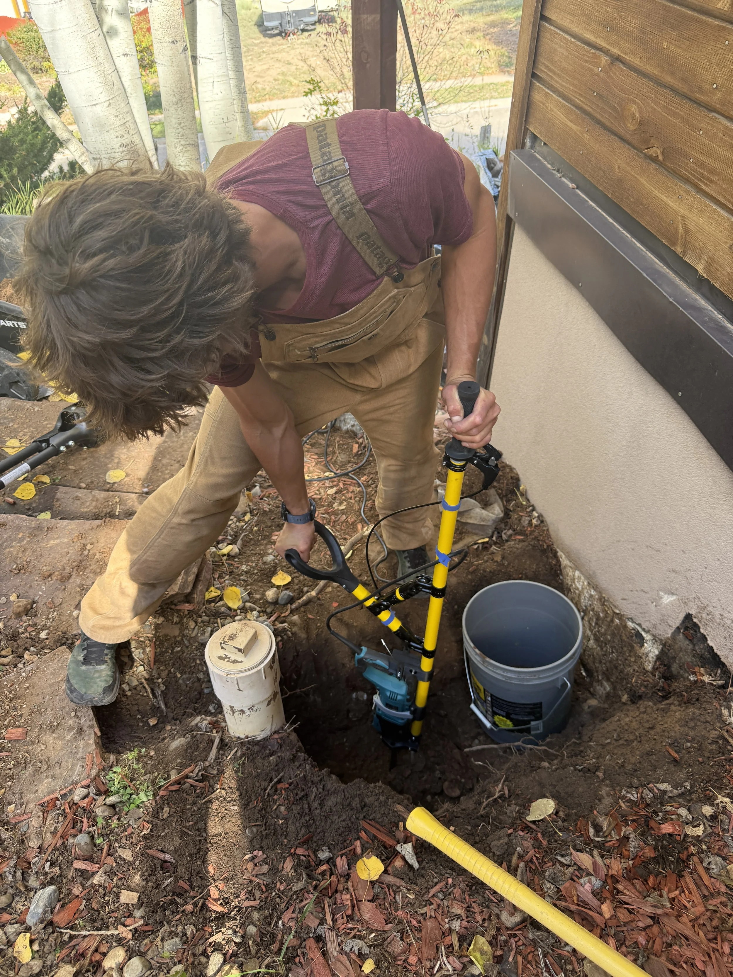 A man is working with a power post-hole digger in a garden or yard area near a house, surrounded by fallen leaves and gardening tools.
