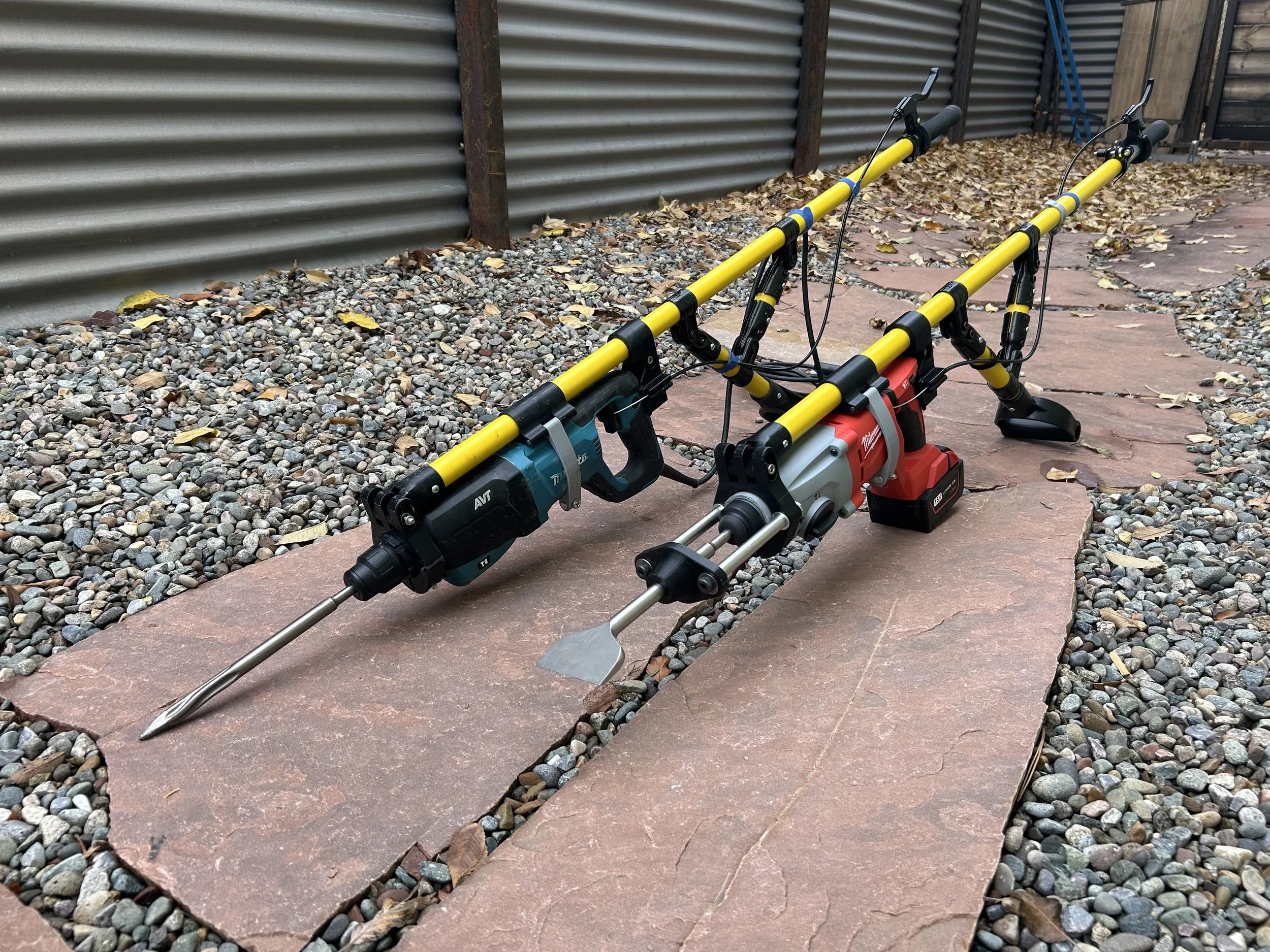 A two-piece power auger drill with yellow handles, a red battery pack, and a metallic drill bit attached, positioned on a flagstone walkway with a gravel border, against a corrugated metal fence.