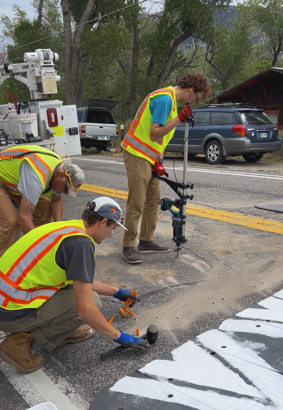 Three construction workers in safety vests working on road repair, with one kneeling and using a hammer, while another stands with a hammer drill, and a third is crouched near the road, with equipment and parked cars in the background.