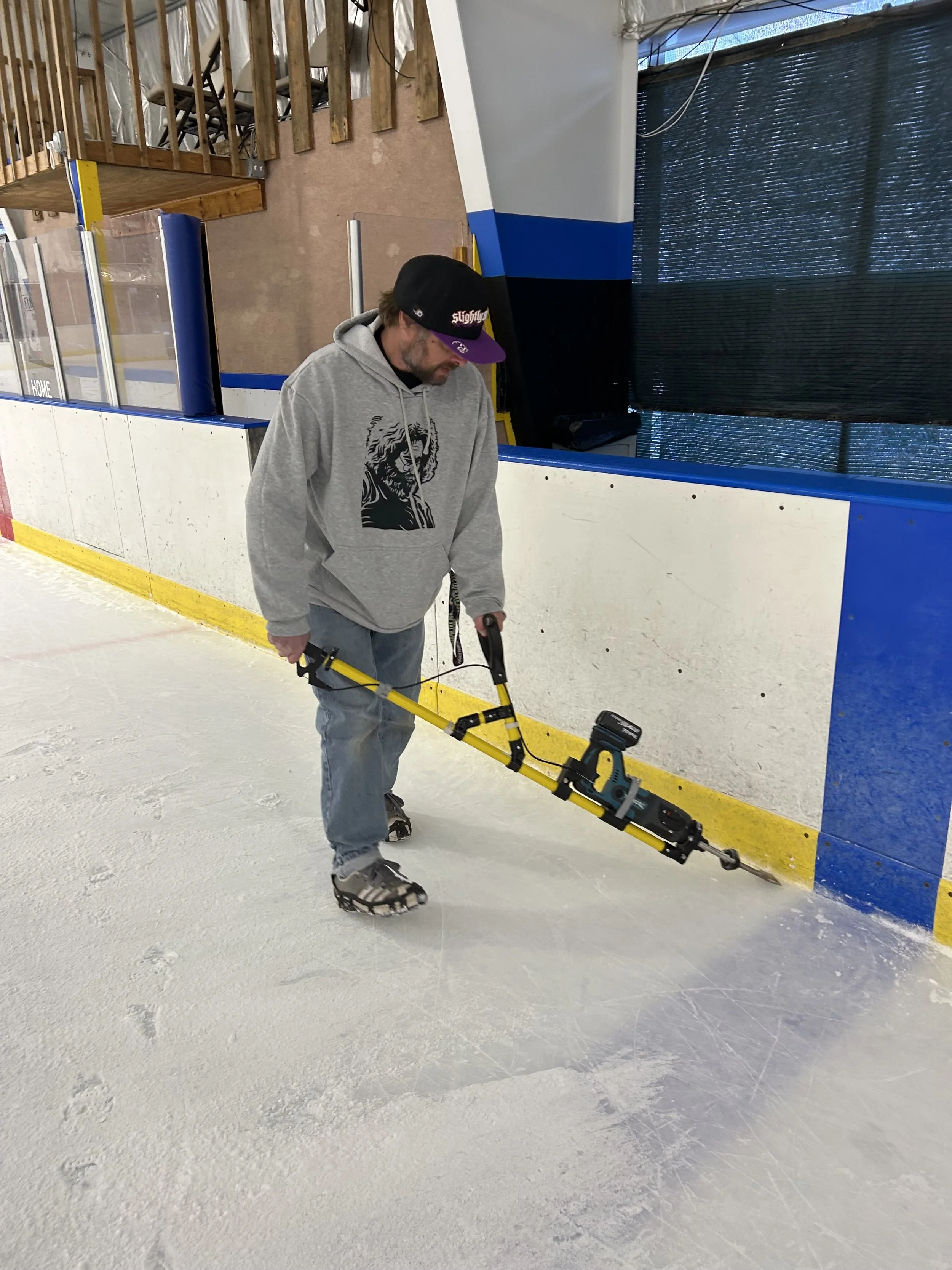 A man is using a rotary hammer drill on an indoor ice hockey rink, wearing a gray hoodie, jeans, and a black cap.