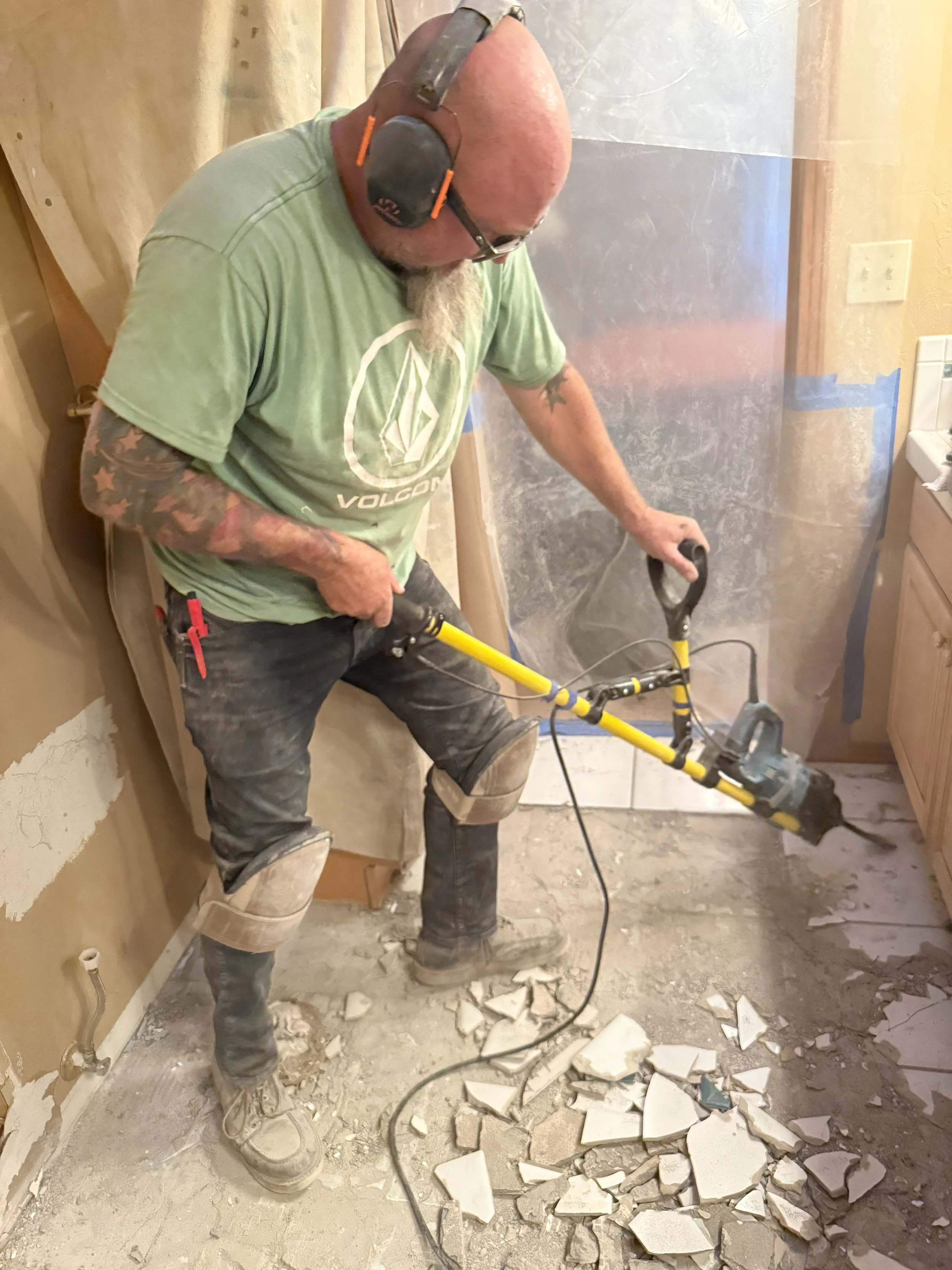 A man using a rotary hammer drill to stand upright and remove tile break up tiles in a bathroom under renovation, surrounded by broken tile pieces on the floor.