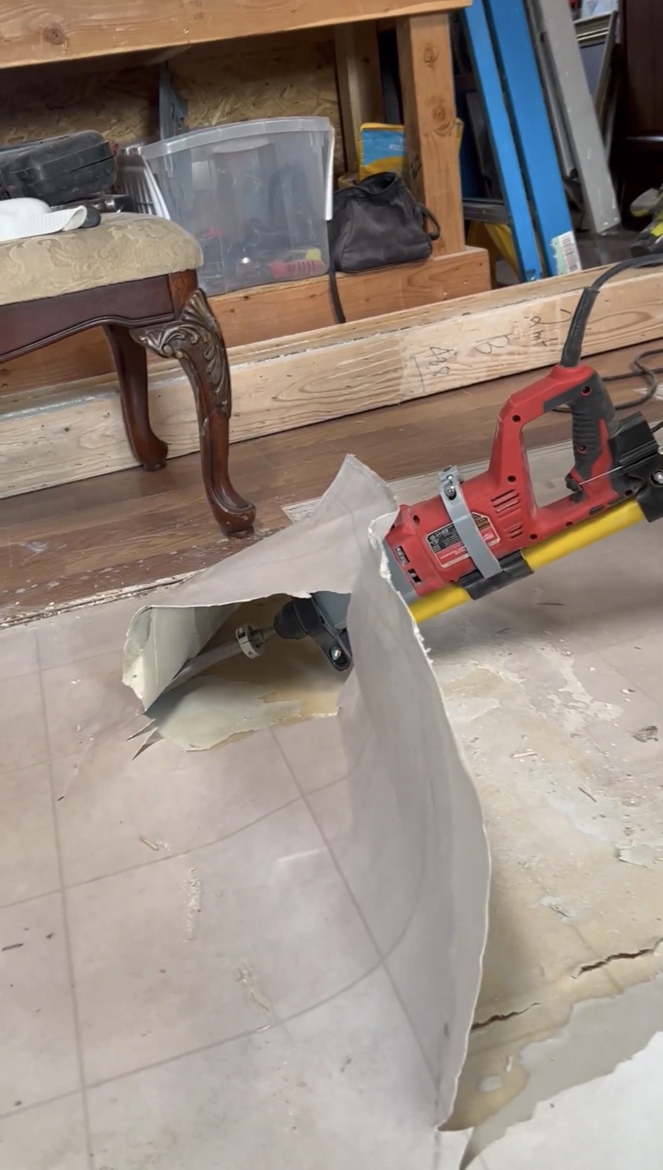 A linoluem floor with a large hole and a red Milwaukee hammer drill demolition drill. A piece of the linoluem is being removed, showing the subfloor underneath.
