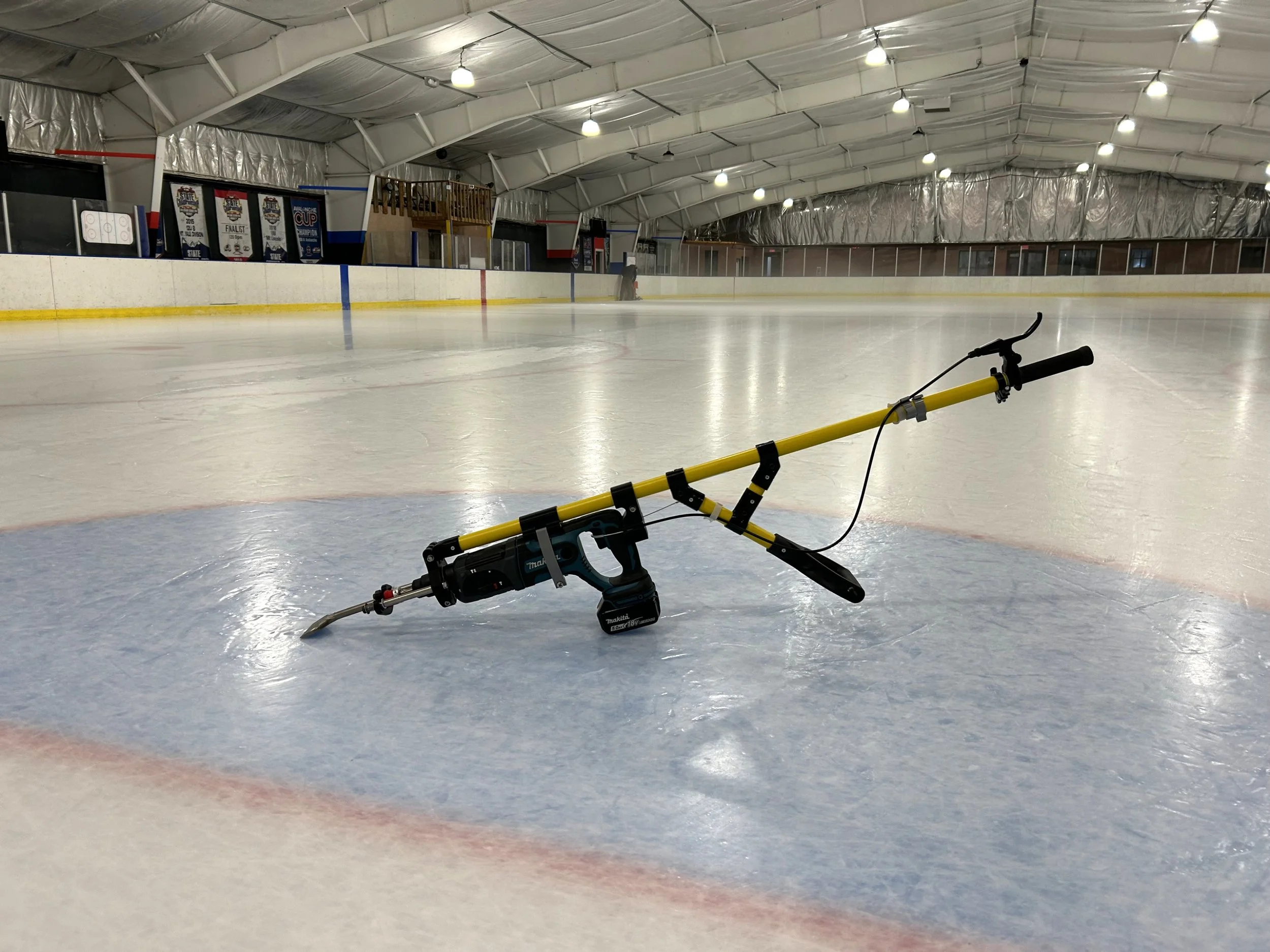 An ice rink with a yellow rotary hammer drill machine resting on the ice. The rink is empty and brightly lit with overhead lights, with banners hanging on the wall in the background.