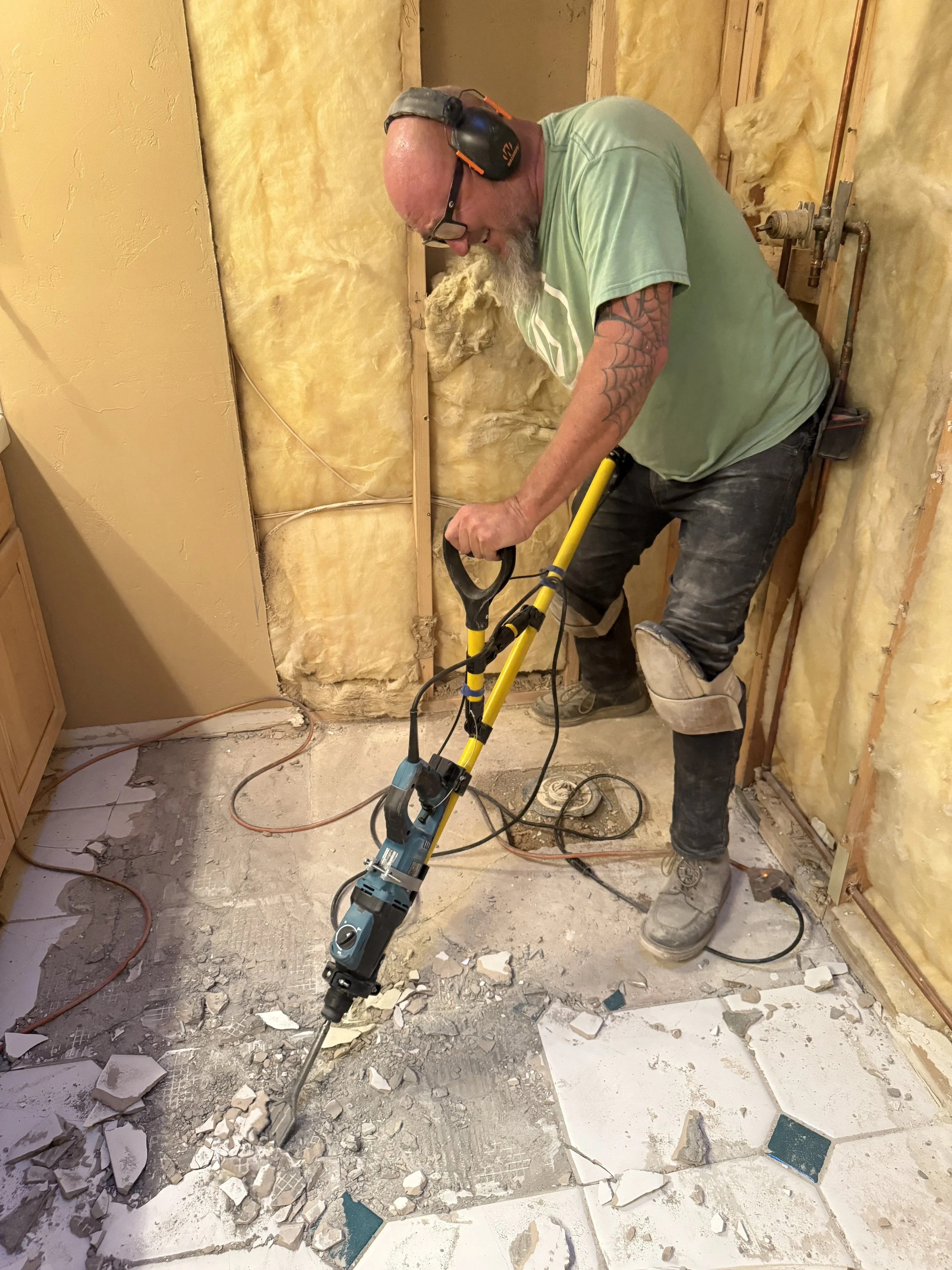 A man with a beard wearing a green shirt, headphones, and glasses uses a rotary hammer drill tool to break up a tile floor in a room under renovation, with yellow insulation and exposed wall studs in the background.