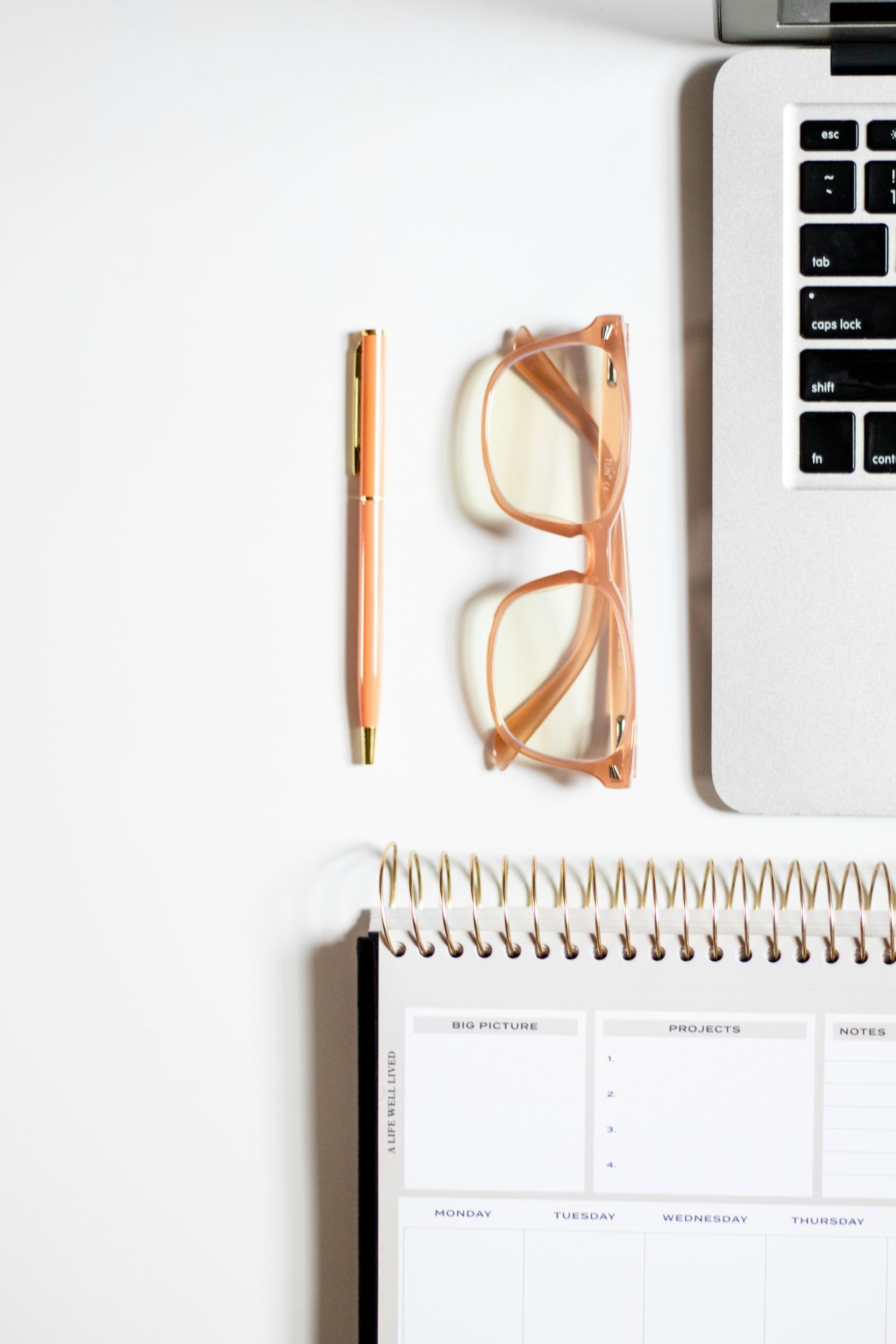 A workspace with a pink pen, pink eyeglasses, a laptop keyboard, and a weekly planner on a white surface.