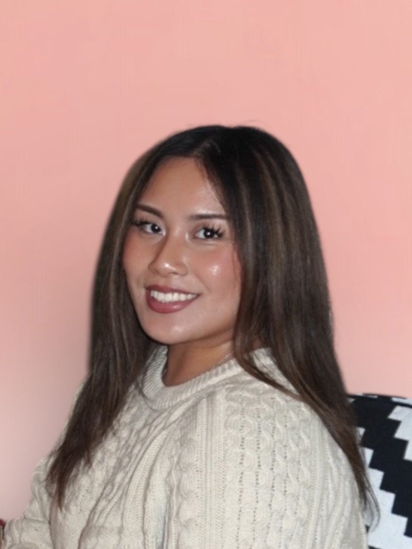 A young woman with long brown hair, wearing a cream knit sweater, smiling, sitting against a pink background.