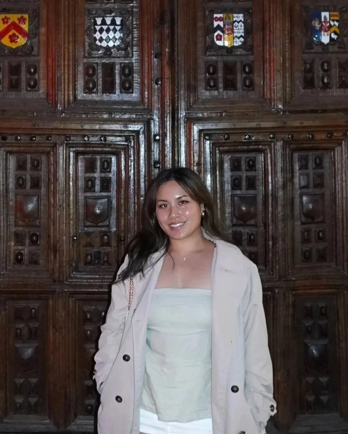 Young woman with long dark hair, wearing a strapless light-colored top and a beige coat, standing in front of a dark wooden panel with carved shields and heraldic imagery.