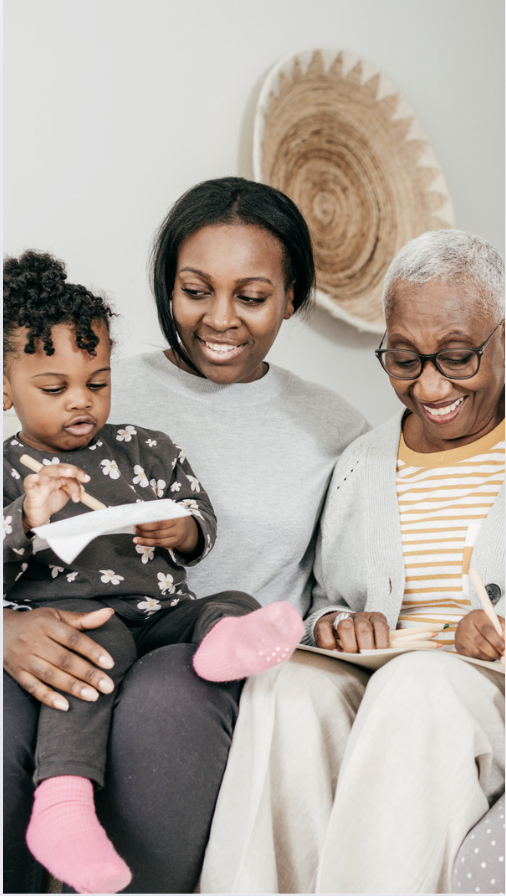 A multigenerational family sitting together, with a young girl drawing on a notepad, a woman smiling at her, and an elderly woman holding a book, in a cozy home setting.