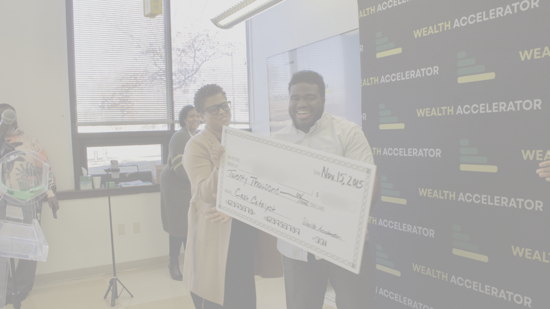 Two people smiling and holding a large check for twenty thousand dollars during a presentation event at Wealth Accelerator. Other individuals are in the background.