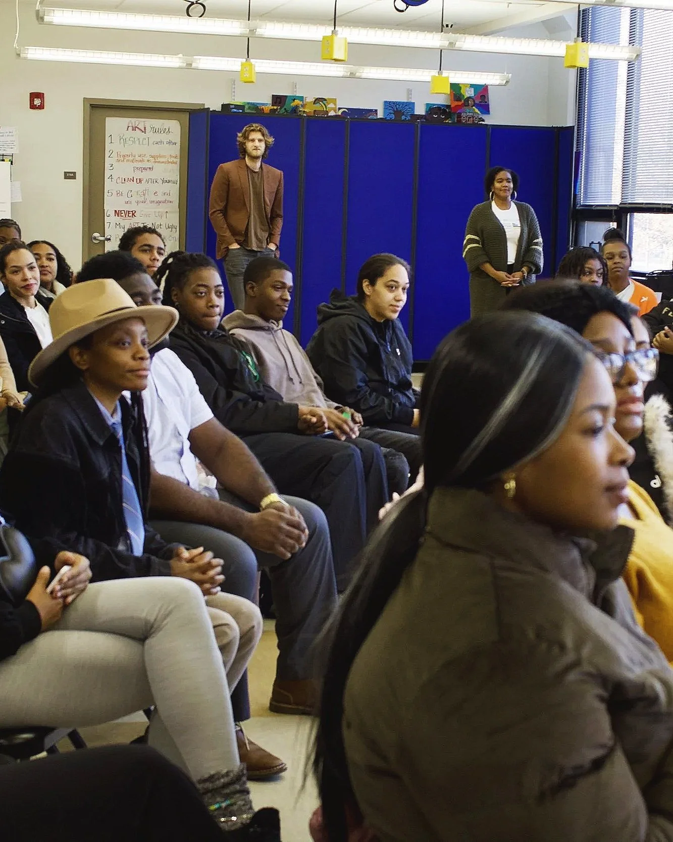 A classroom filled with diverse students seated and listening, with teachers standing at the back, a whiteboard with rules, and colorful artwork on the walls.