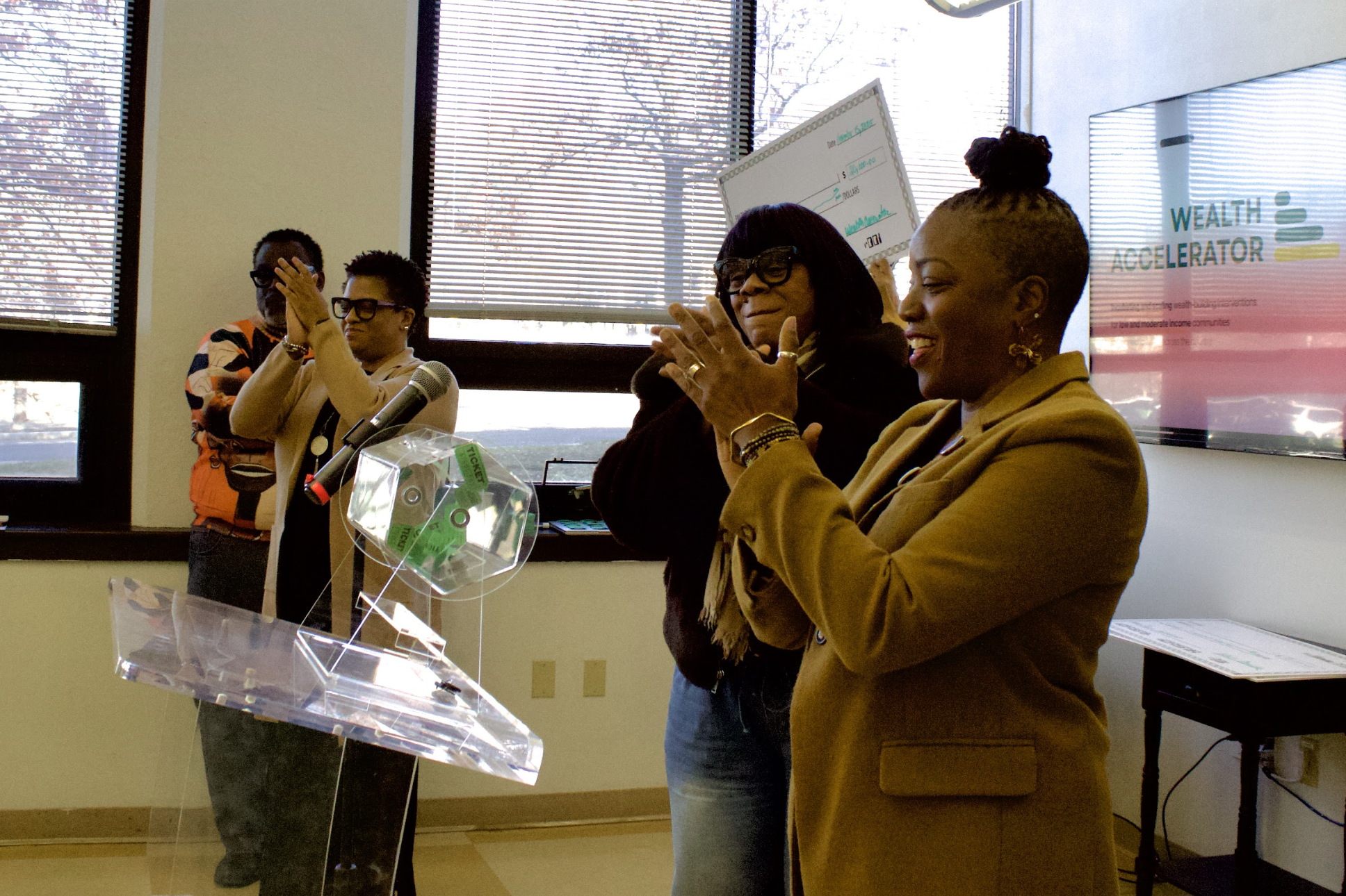 Group of four diverse women standing inside a room, clapping and smiling during an event. One woman is at a clear podium, and a digital screen displays 'WEALTH ACCELERATOR' in the background.