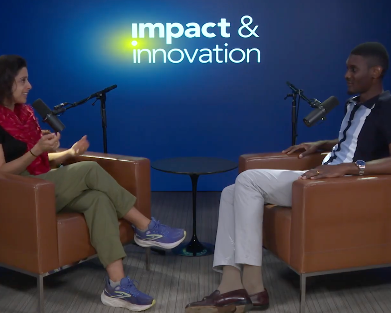 Two people engaged in a podcast or interview, seated in armchairs facing each other with microphones, in front of a blue background displaying the words 'impact & innovation'.