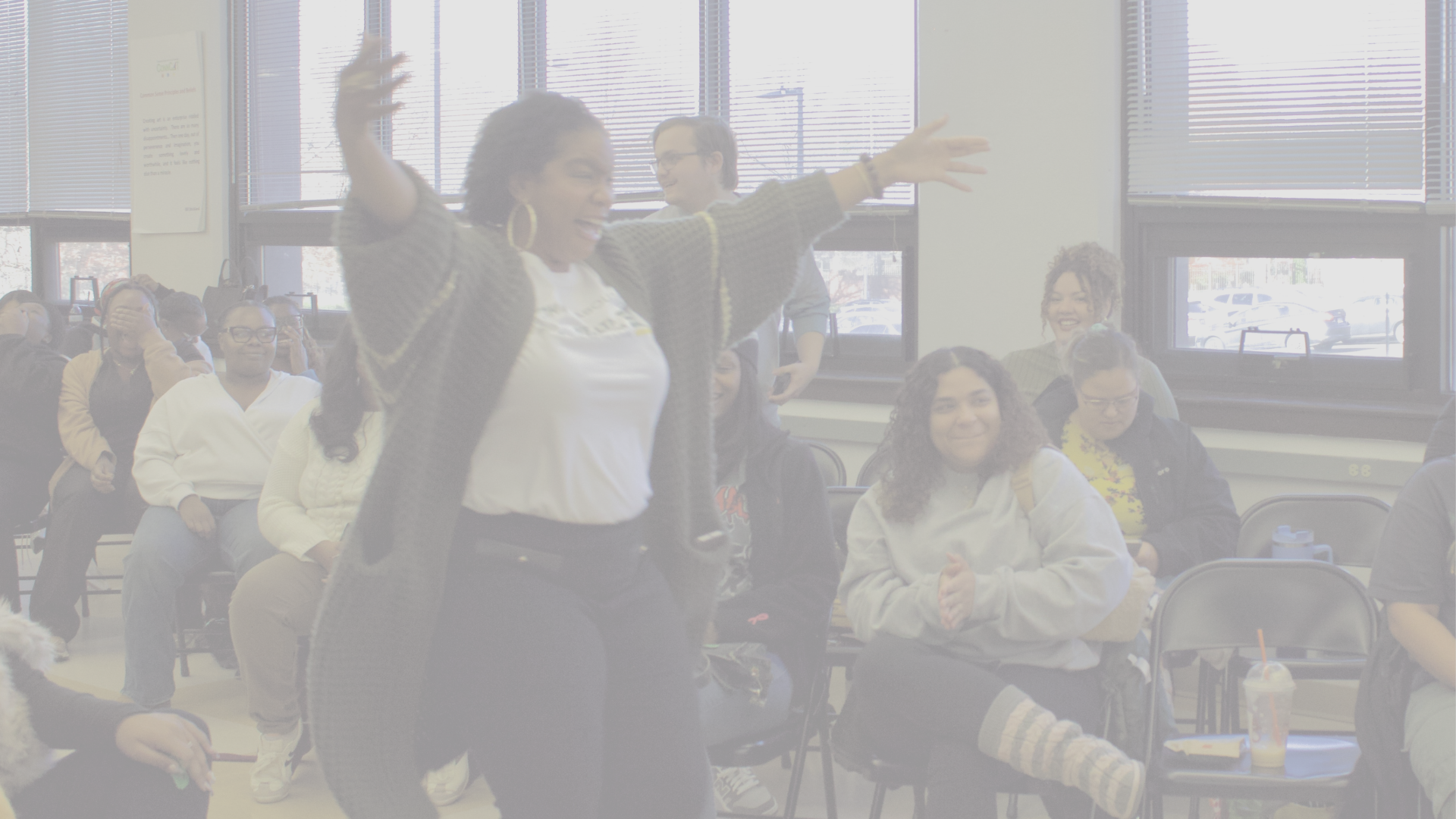A woman with curly hair and large hoop earrings is standing with her arms raised and smiling in a classroom or workshop setting. Several people are sitting and standing around her, some clapping and smiling, with large windows in the background allowing natural light inside.