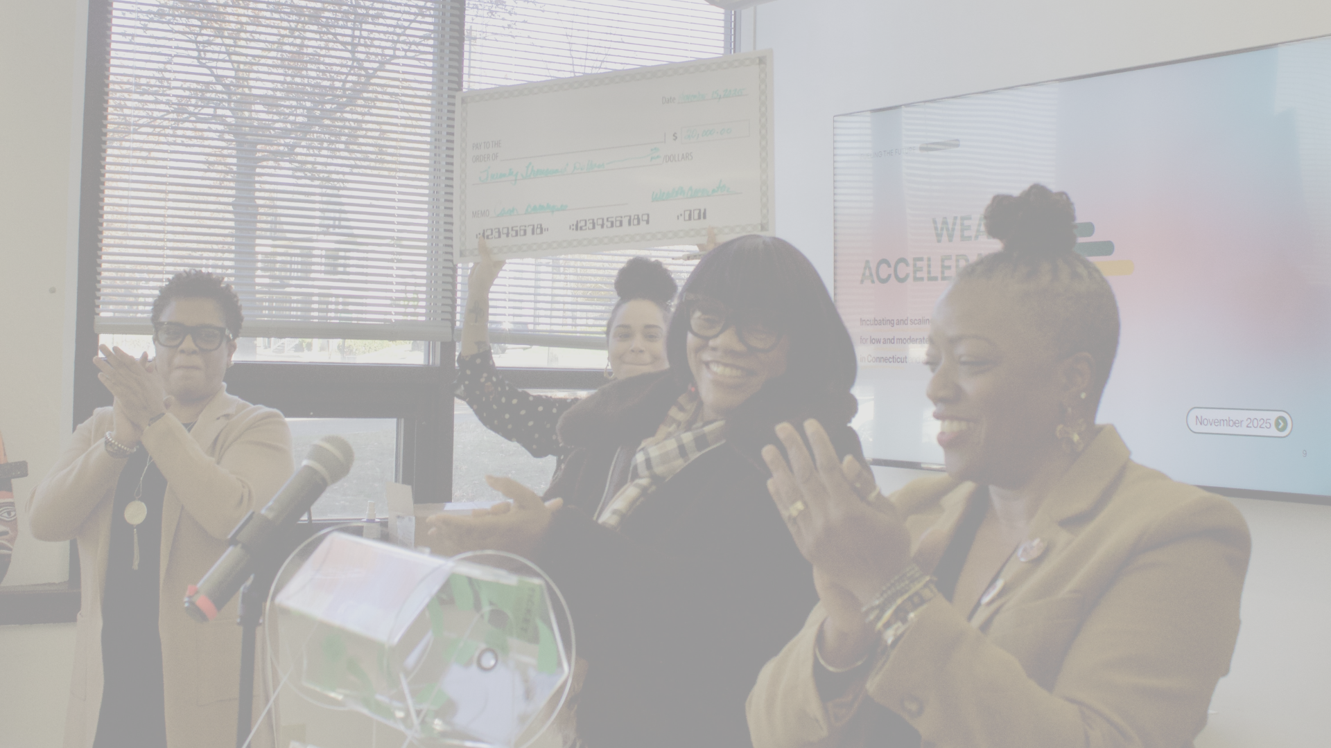 Three women and a man celebrating an achievement in a meeting room, with one woman holding a large check for $20,000, and a presentation screen displaying 'WEAR ACCELERATE' and a date of November 2025.