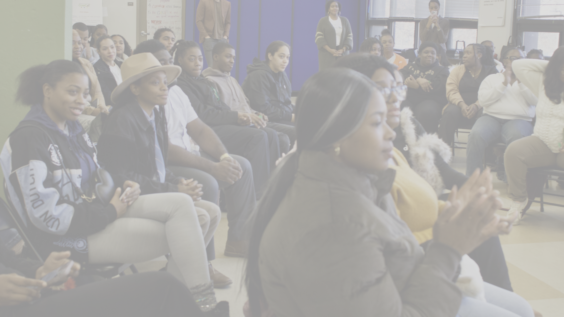 A diverse group of people sitting and standing in a classroom or meeting room, attentively listening to someone outside the frame.