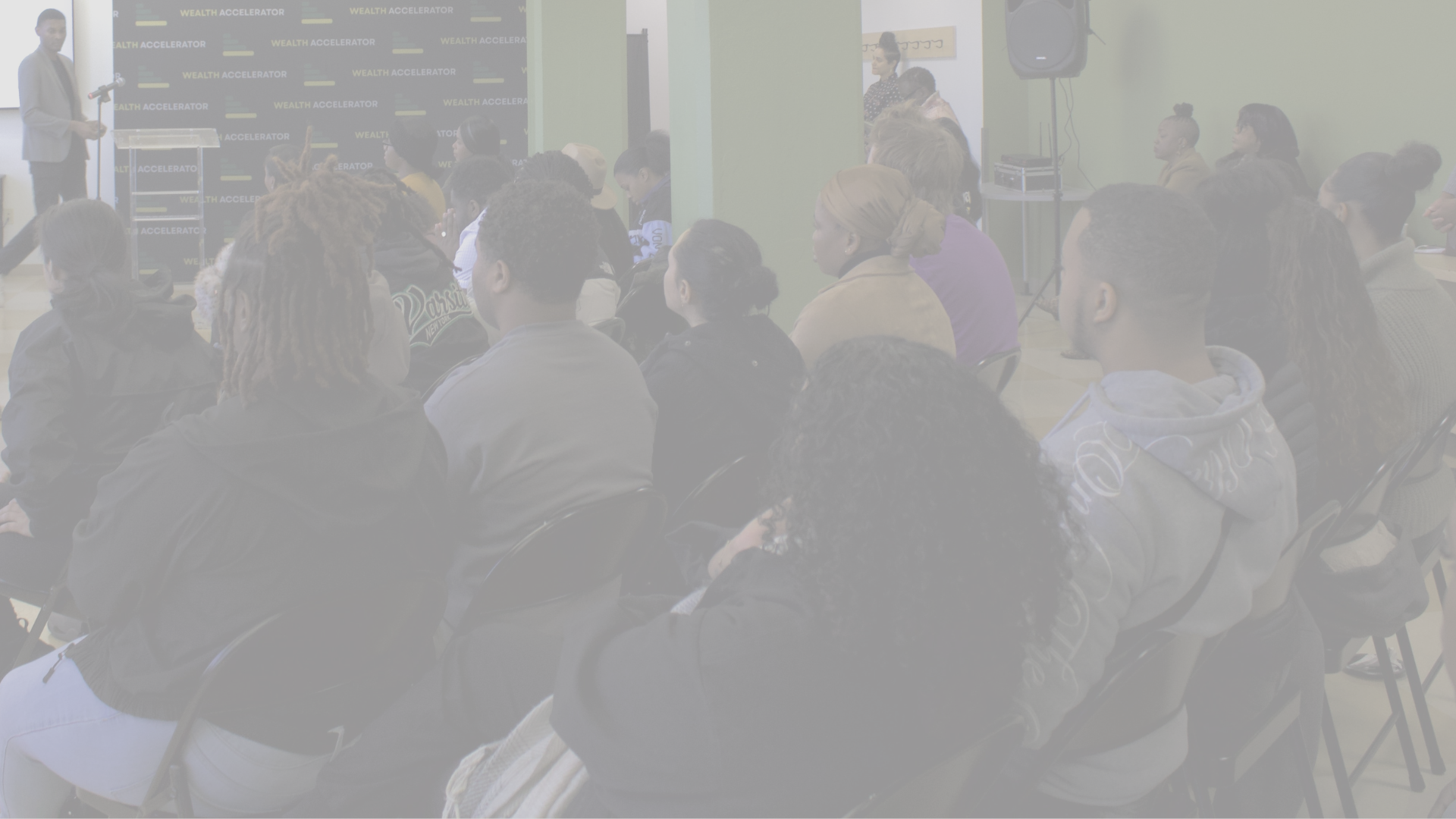 A group of people attending a presentation or seminar with a speaker at the front near a backdrop labeled 'Wealth Accelerator', seated in a room with light green walls.