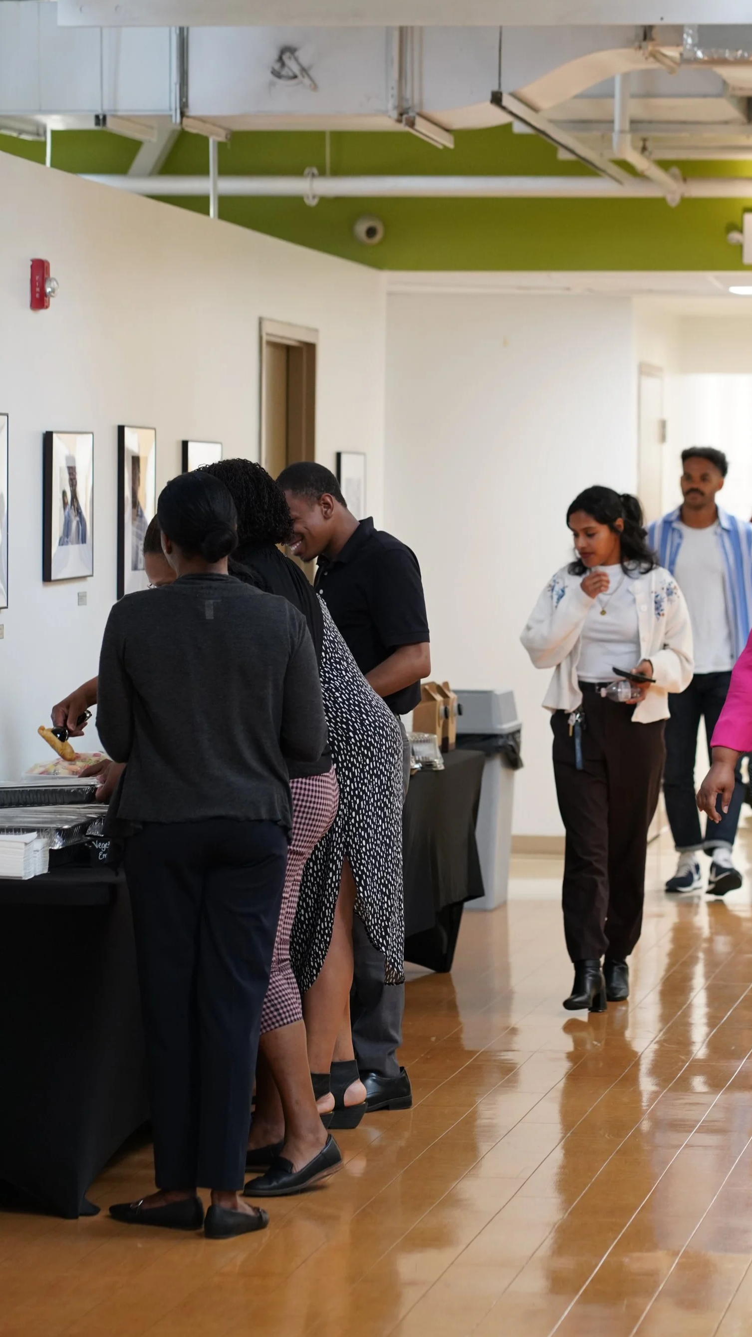 People gathering around a buffet table at an art gallery or event, with framed artwork on the walls and a green ceiling.