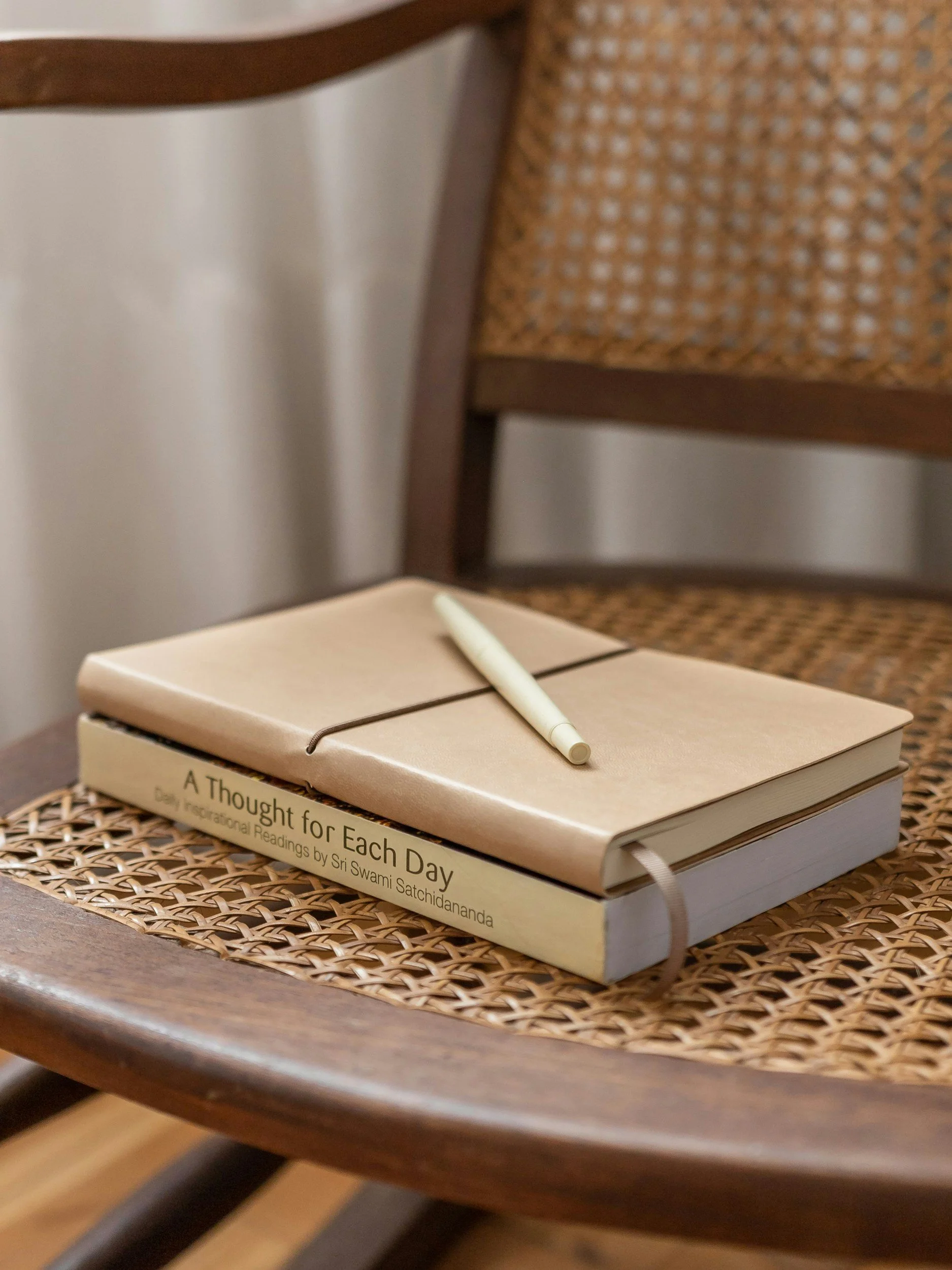 A closed beige notebook with a matching pen resting on top, placed on a woven placemat on a wooden chair. The book is titled "A Thought for Each Day" by Sri Swami Satchidananda.