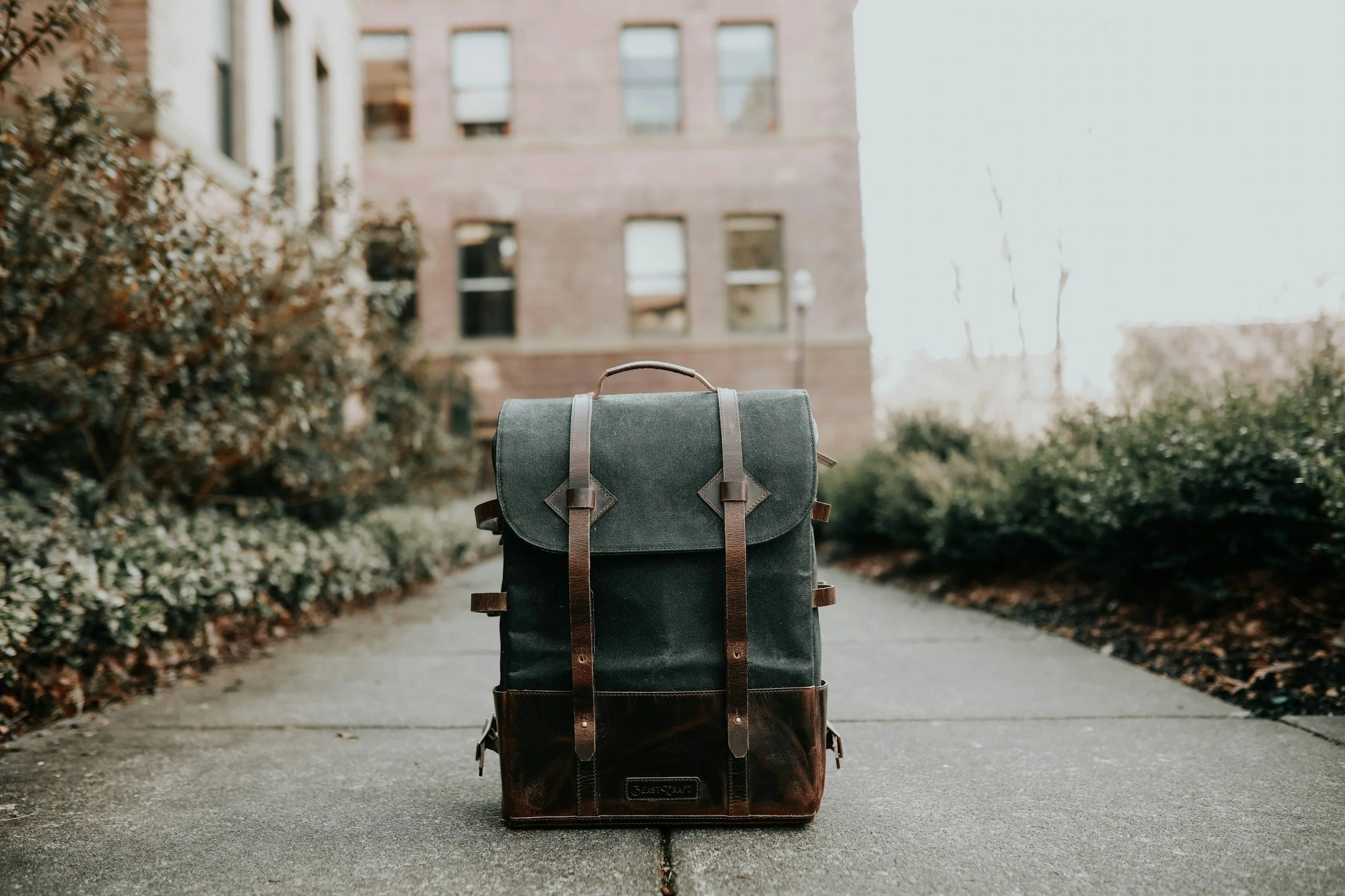 A backpack with a vintage style, featuring a dark canvas body and leather straps, placed on a sidewalk outdoors with buildings and bushes in the background.