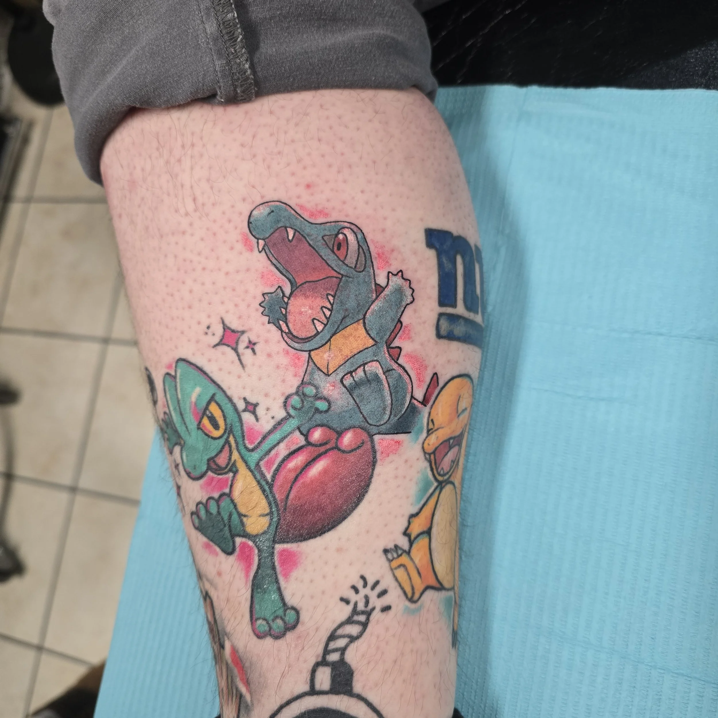 Colorful tattoo of three Pokémon characters, including Totodile, Charmander, and Squirtle, on a person's thigh.