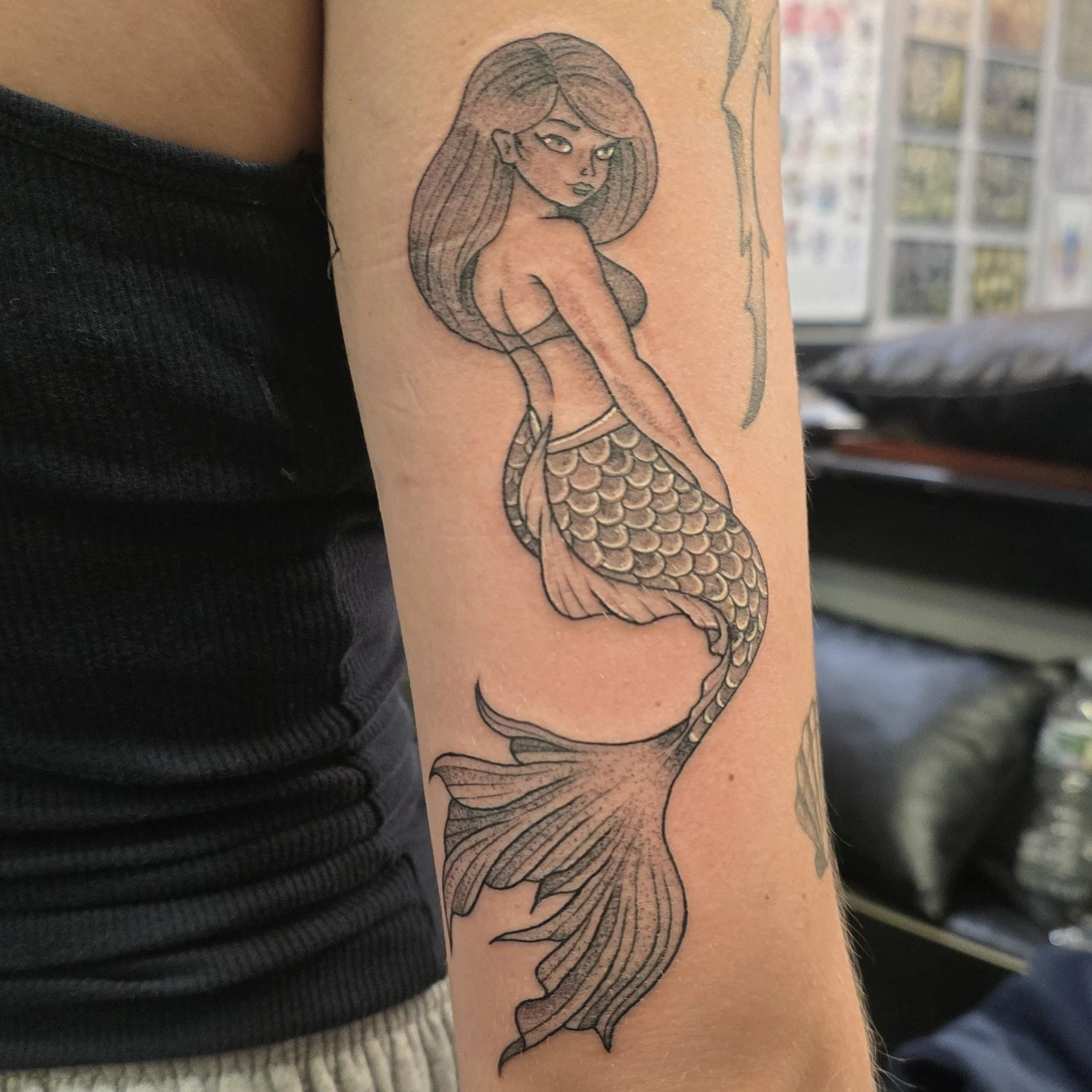 Tattoo of a cartoon-style mermaid with long hair and a scale-covered tail, facing sideways with her head slightly turned.