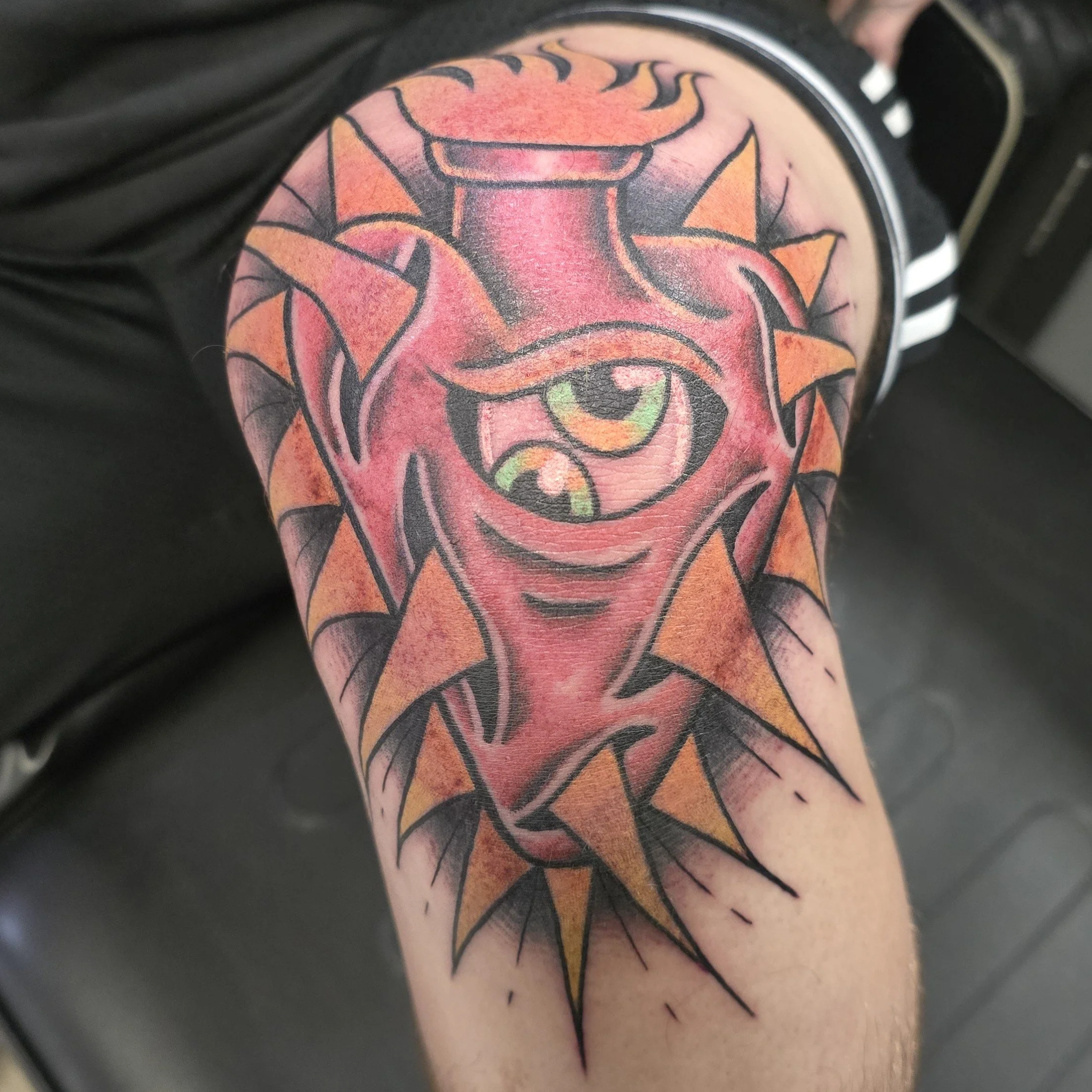 Tattoo of a one-eyed monster with sharp orange teeth, surrounded by orange and black spiked elements.