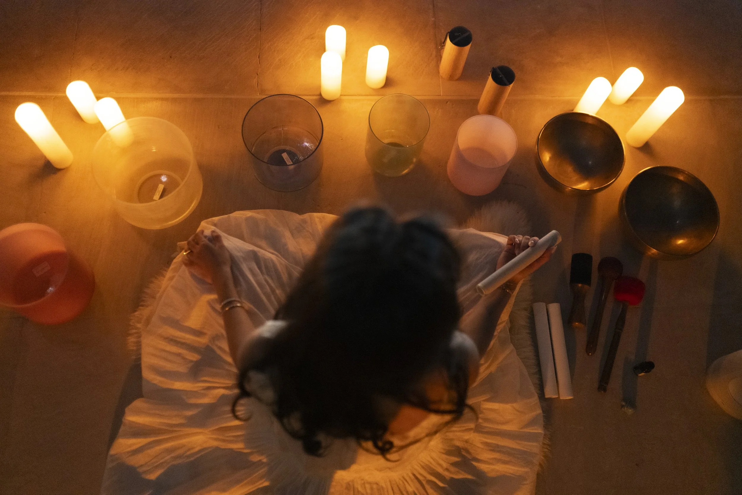 A woman sitting cross-legged on a chair surrounded by lit candles and singing bowls, viewed from above.