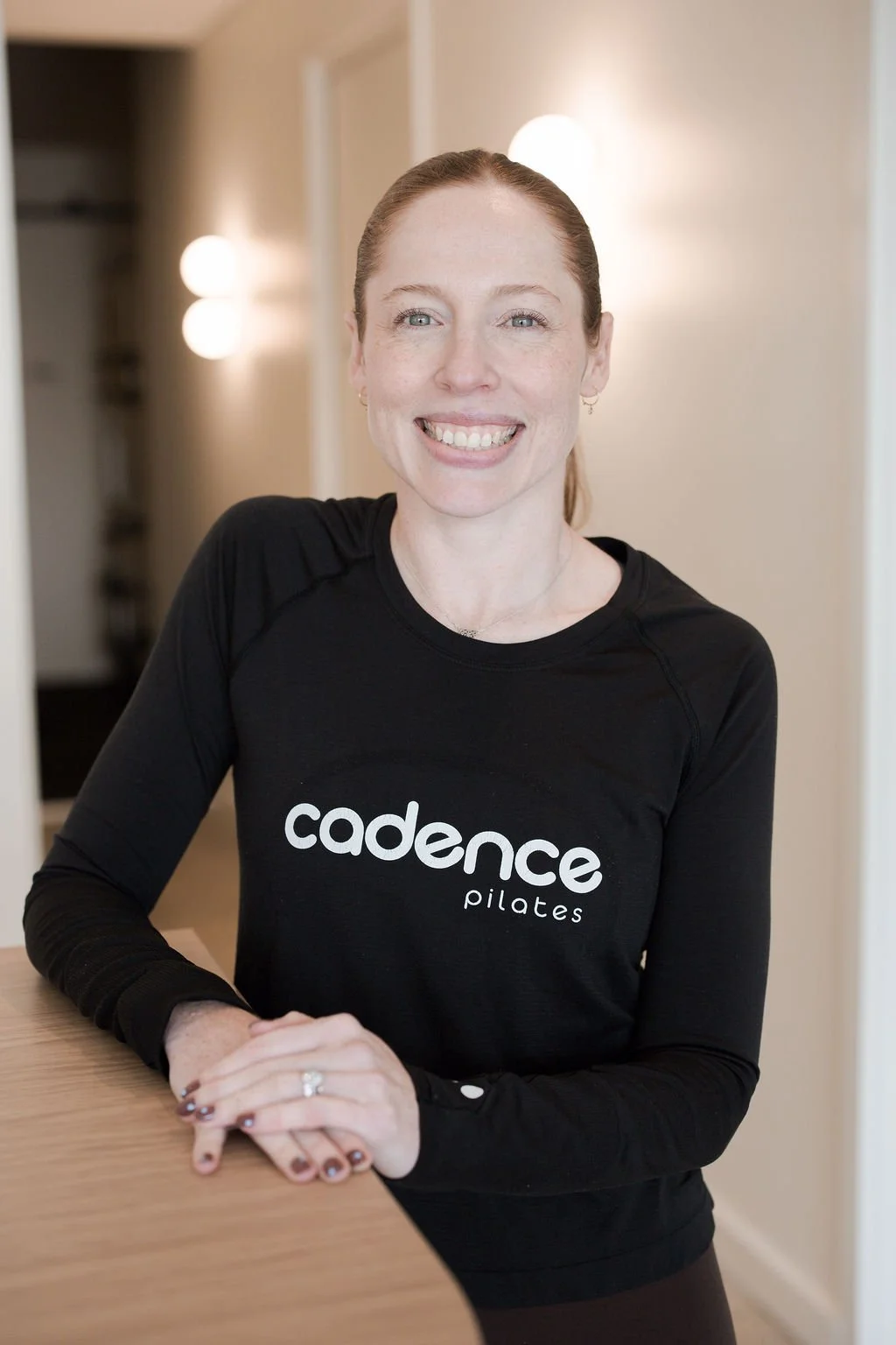 A woman with red hair smiling, wearing a black sleeveless top with 'Cadence Pilates' logo, sitting against a light-colored wall.