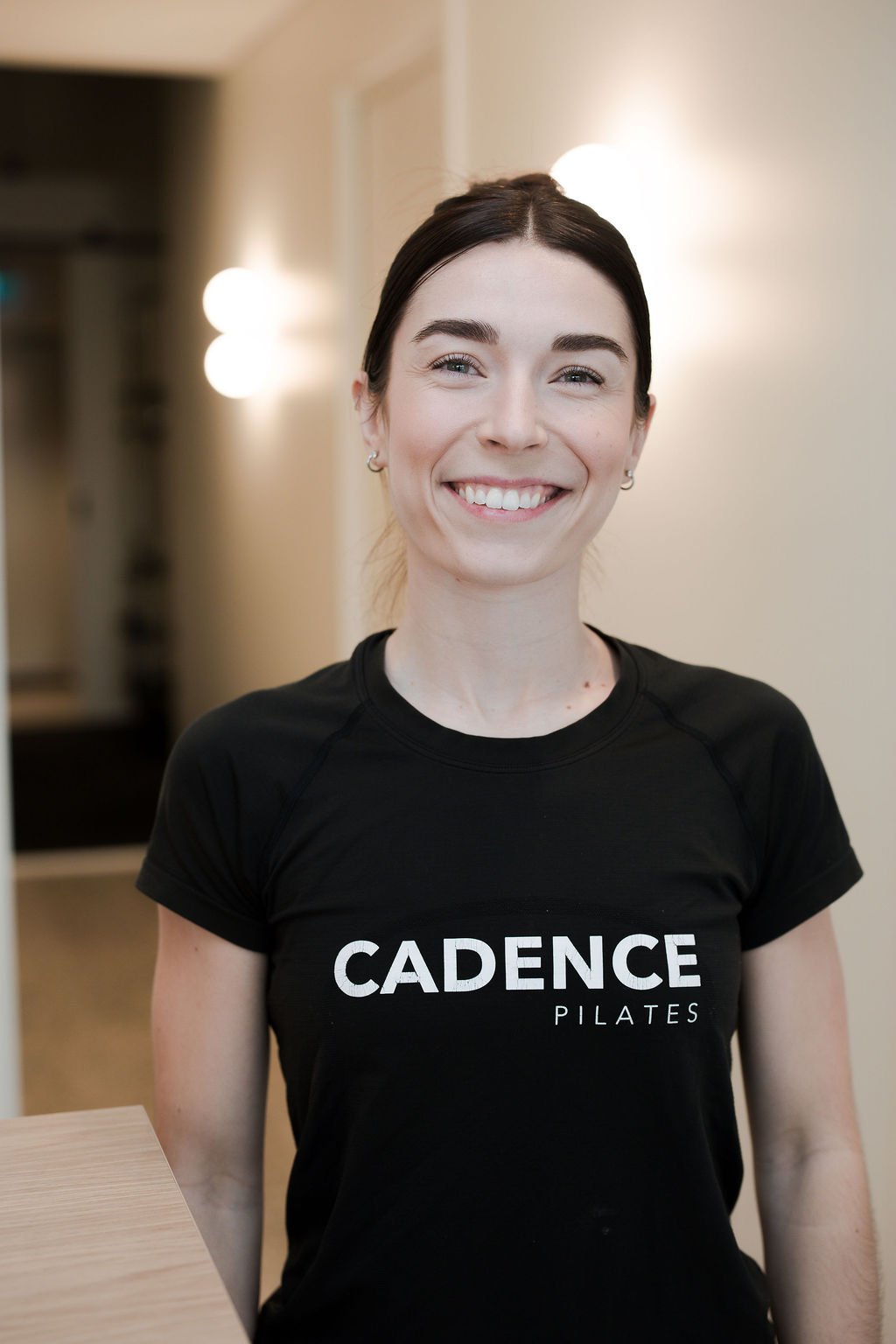 Smiling young woman with short brown hair, wearing a black t-shirt with 'cadence pilates' on it, standing in front of a wooden-paneled wall with warm lighting.