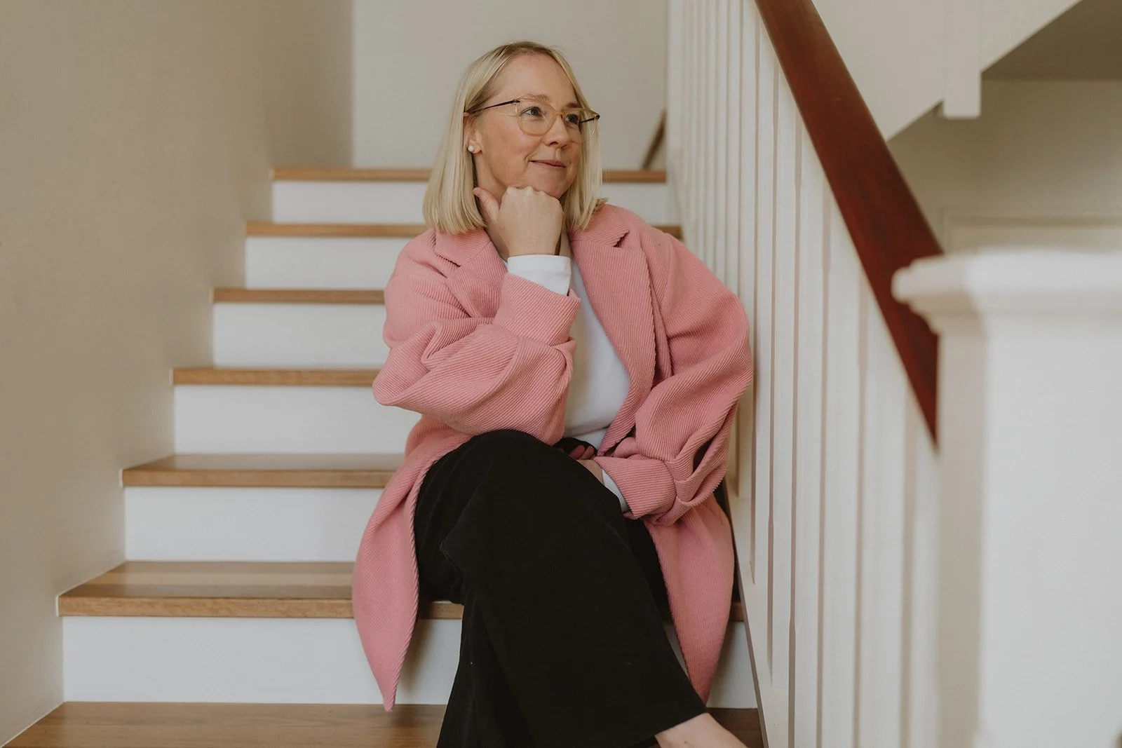 A woman sitting on wooden stairs with her elbow resting on her knee, dressed in a pink blazer, white shirt, and black pants, looking thoughtfully to the side.