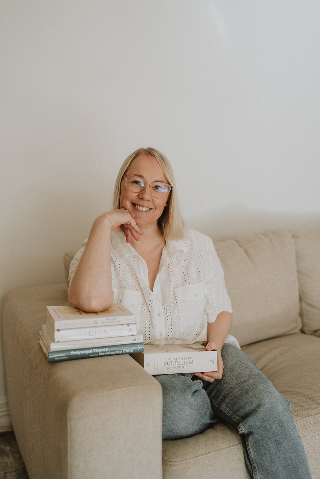 A woman with blonde hair and glasses sitting on a beige sofa, smiling at the camera, with a stack of books on the armrest and holding a book titled 'The Complete Buddhism for Mothers'.