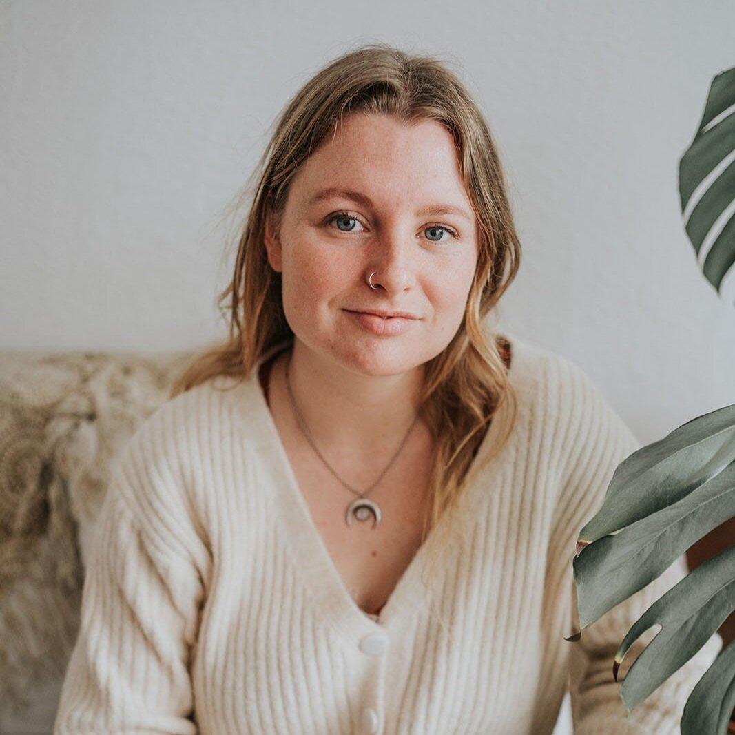 A young woman with light brown hair, blue eyes, and a nose piercing, wearing a cream-colored cardigan and a necklace with a crescent moon pendant, sitting on a beige couch with a large green plant nearby.
