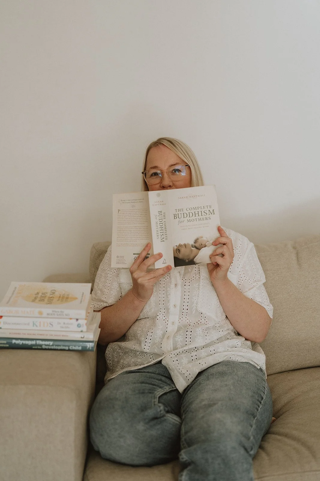 A woman with blond hair and glasses sitting on a beige couch, holding a book titled 'The Complete Buddhism for Mothers' in front of her face. A stack of books is on the armrest of the couch.