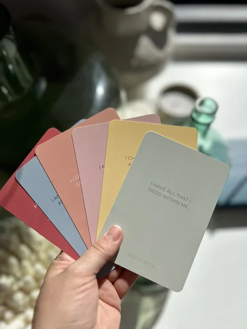 Person holding colorful cards with affirmations, including "I have all that I need within me," in front of a blurred background with a window and various objects.