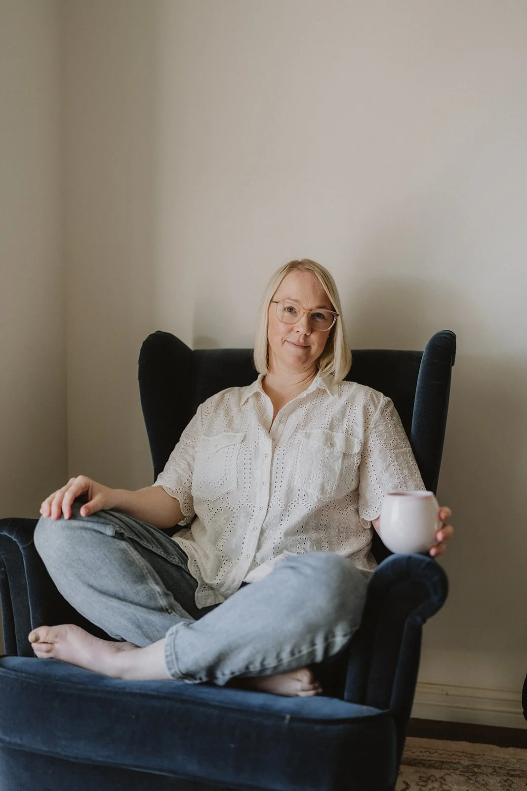 A woman with blonde hair, glasses, and a white eyelet blouse sitting cross-legged in a black armchair, holding a mug in her right hand inside a room with plain walls.