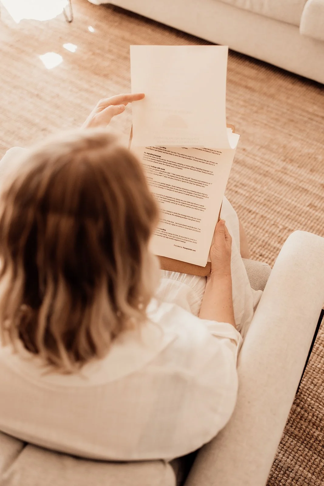 A person with shoulder-length hair is sitting on a beige upholstered chair, reading a stack of papers or documents in a room with a wooden floor and a beige couch.