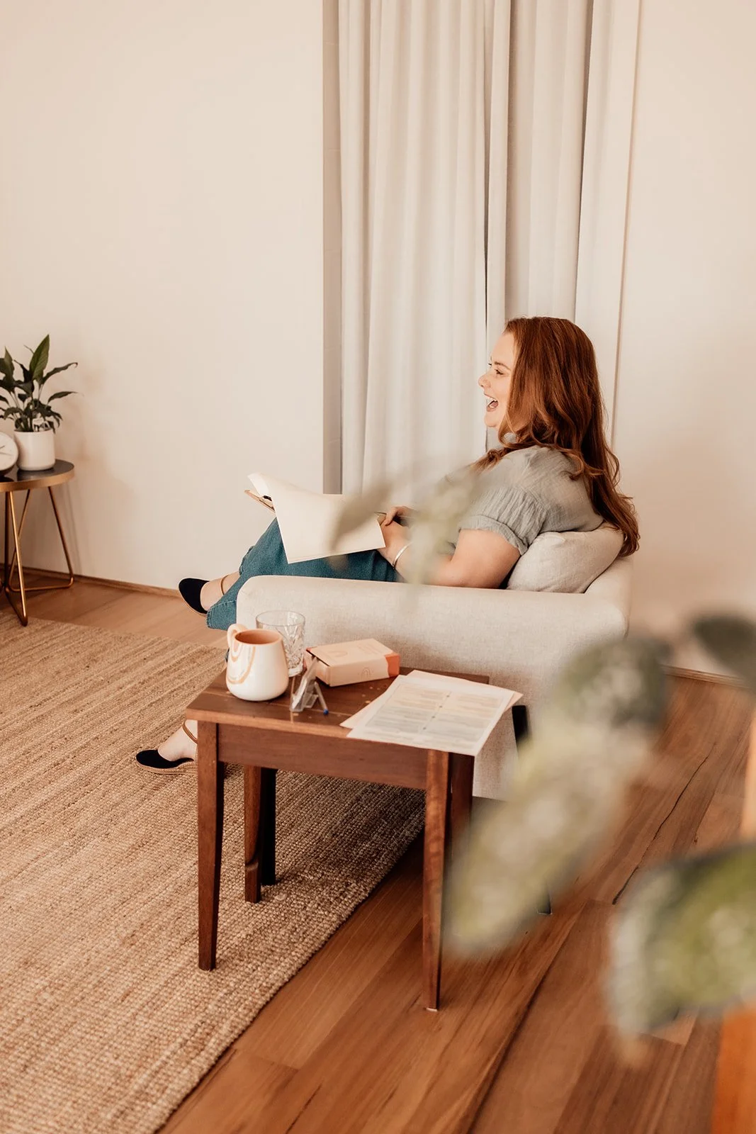A woman with red hair sitting on a beige couch, laughing and reading a book in a cozy living room with a wooden floor, a small wooden side table with a jug, glass, and papers, and a plant on a stand.