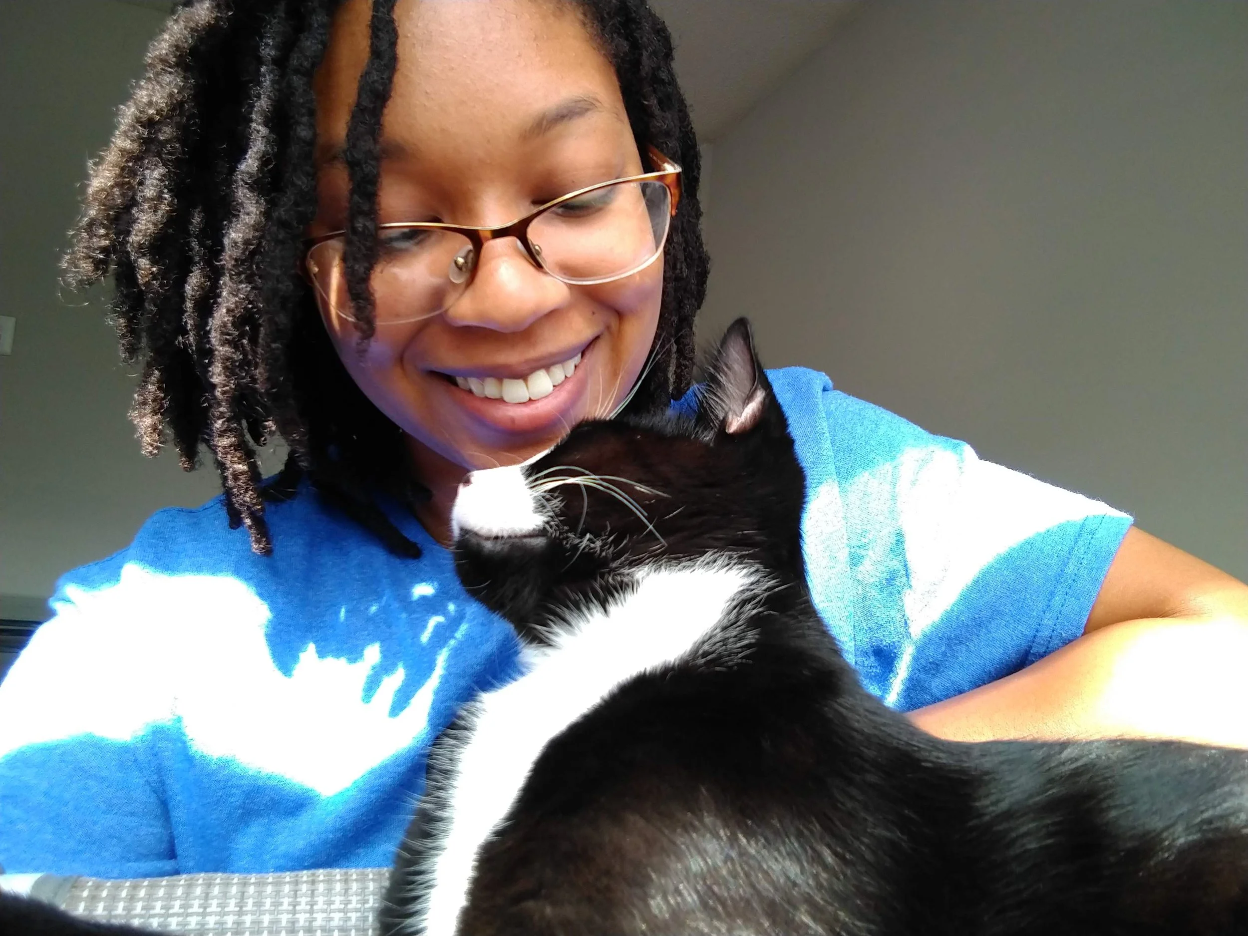 A woman with glasses and braids smiling as she cuddles a black and white cat.