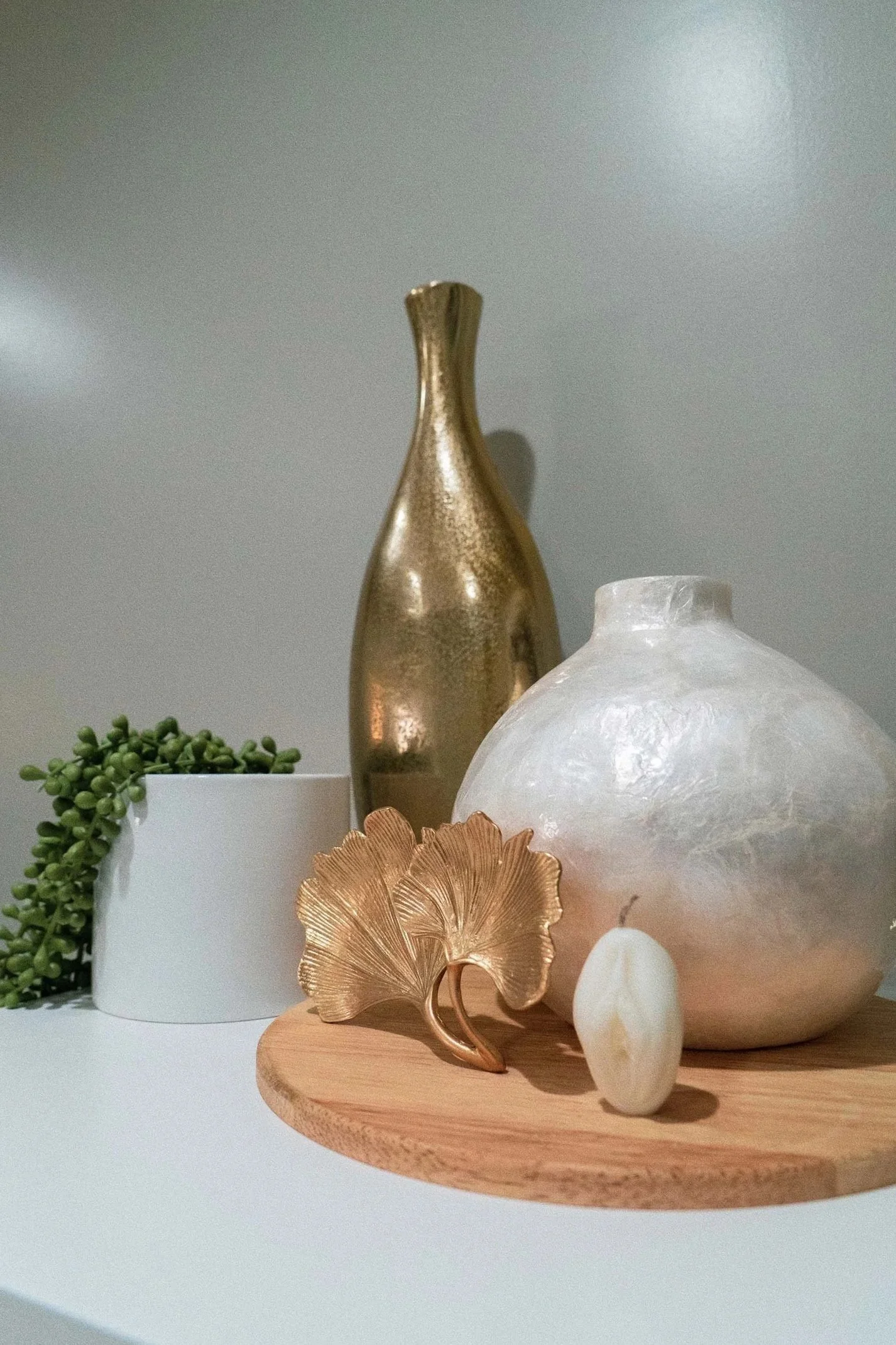 Decorative vases and objects on a wooden tray, including a gold metallic vase, a large white marble-like vase, a seashell-shaped gold ornament, and a small white stone, with some green plant spilling from a white pot.