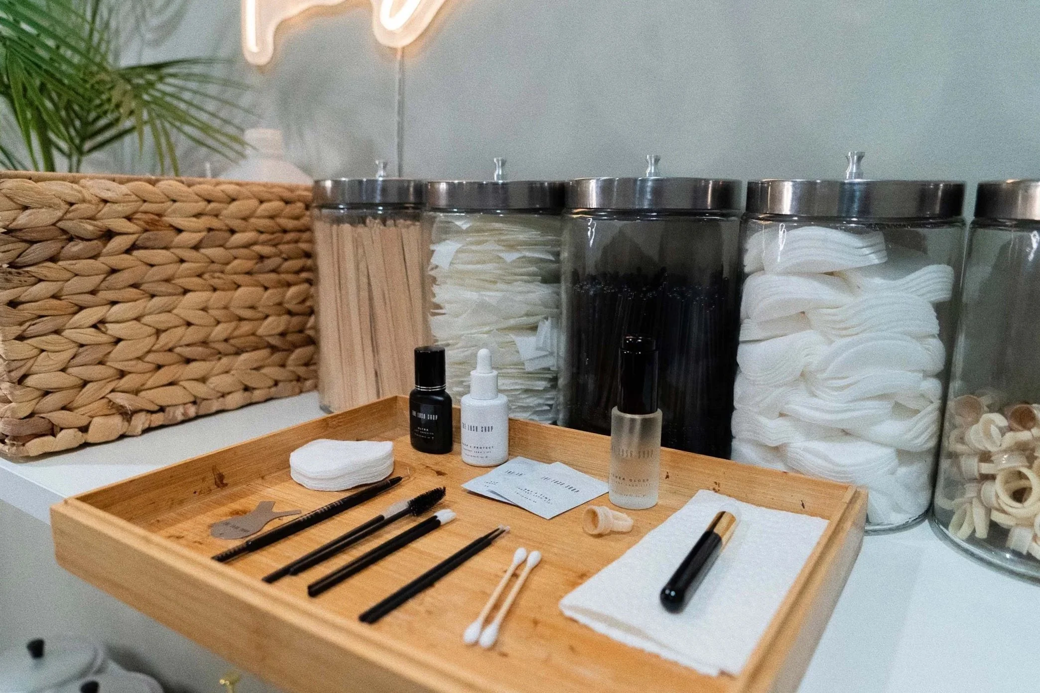 A collection of skincare or beauty tools and accessories on a wooden tray, with jars of cotton pads and swabs in the background.