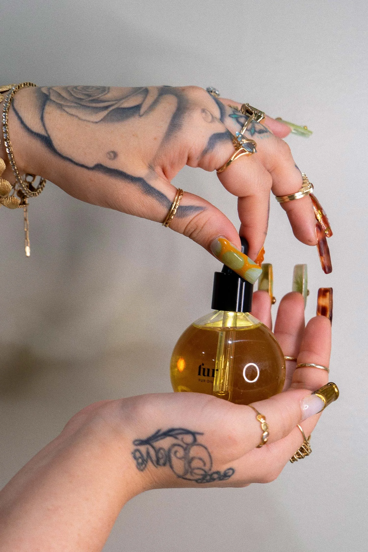 Two hands with multiple rings, bracelets, and tattoos are holding a round oil bottle with a black cap.
