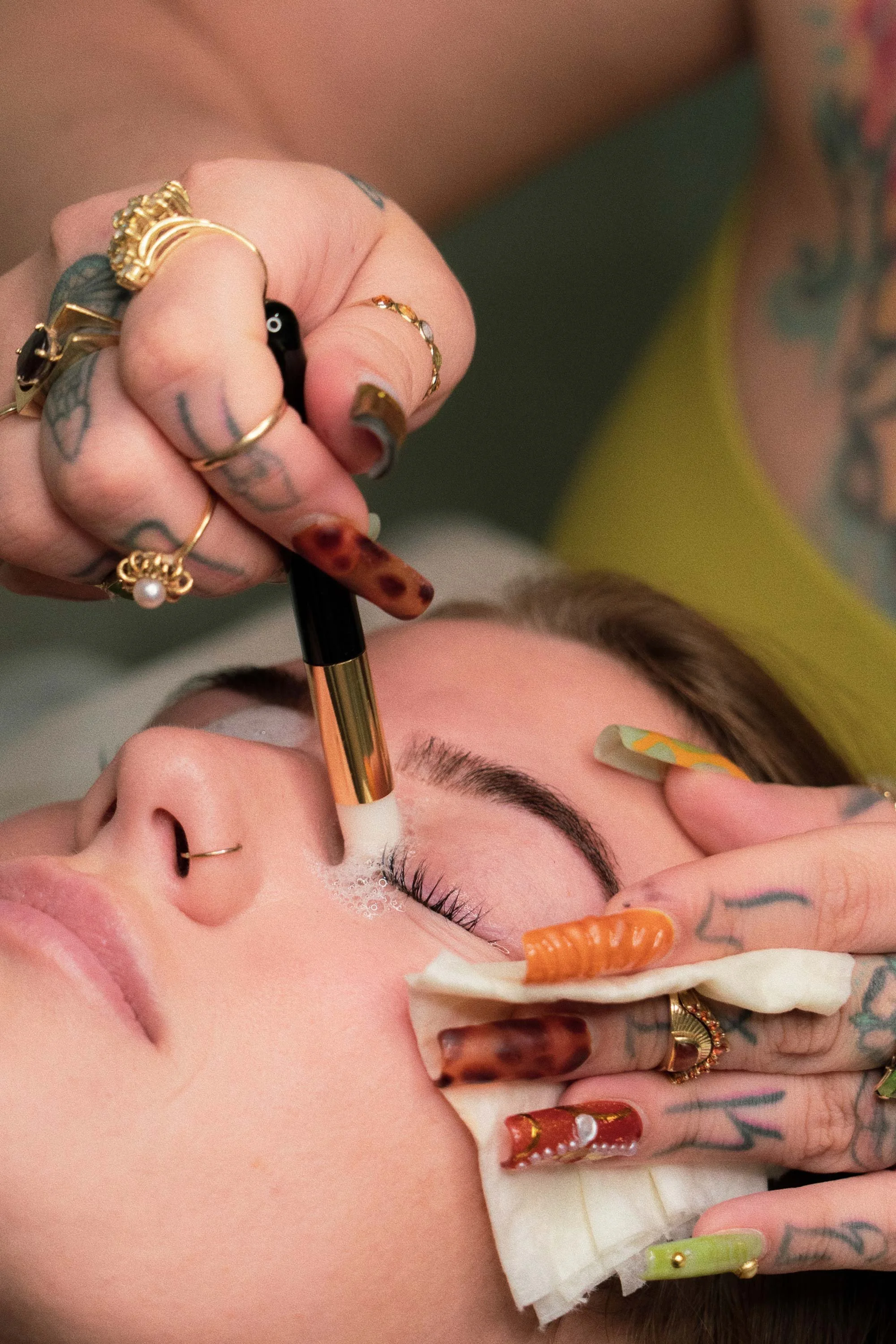 A person with tattoos and multiple rings getting a facial treatment, with a cotton pad held by another person.