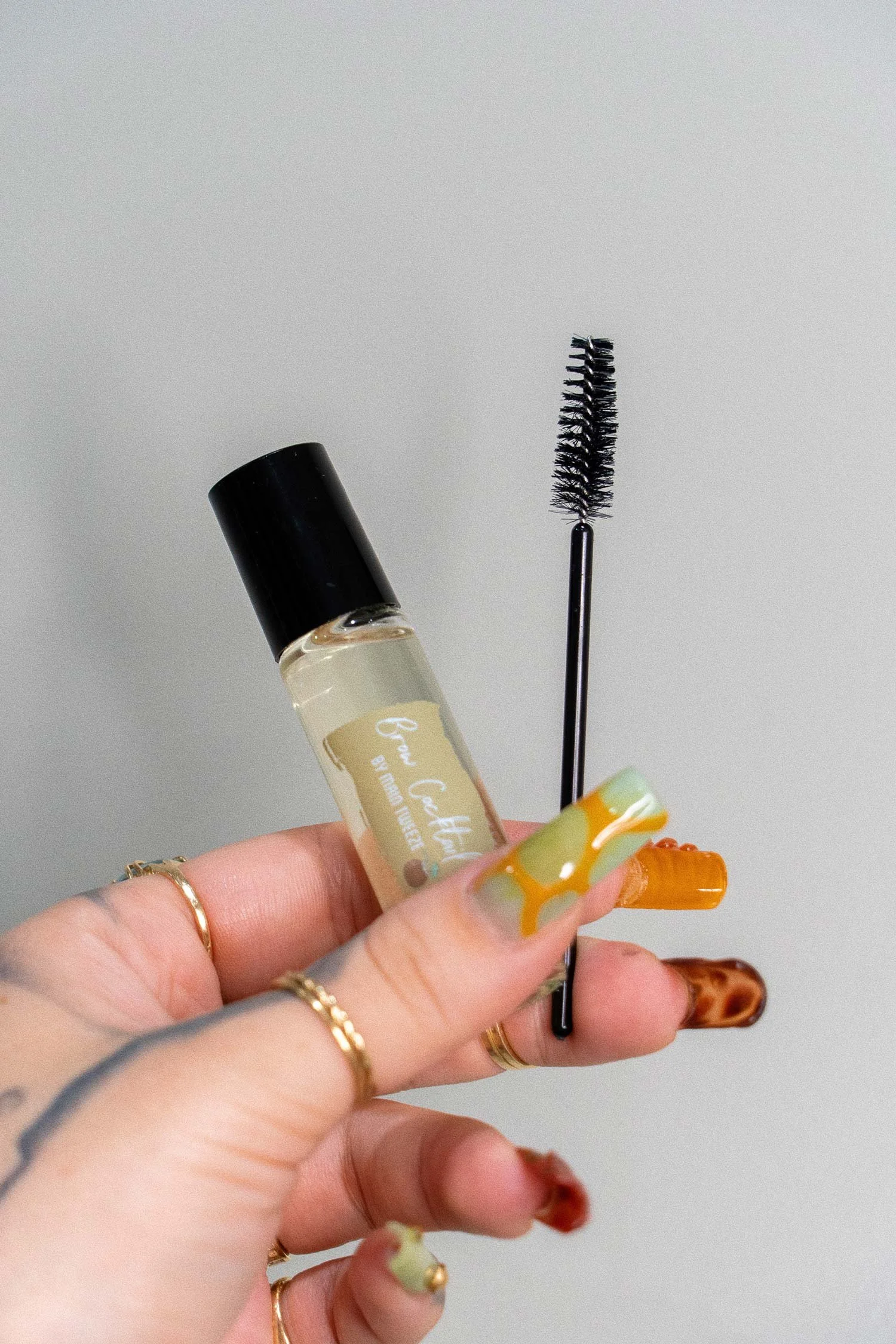 Hand holding a small bottle of lash product and a mascara wand with a grey background.