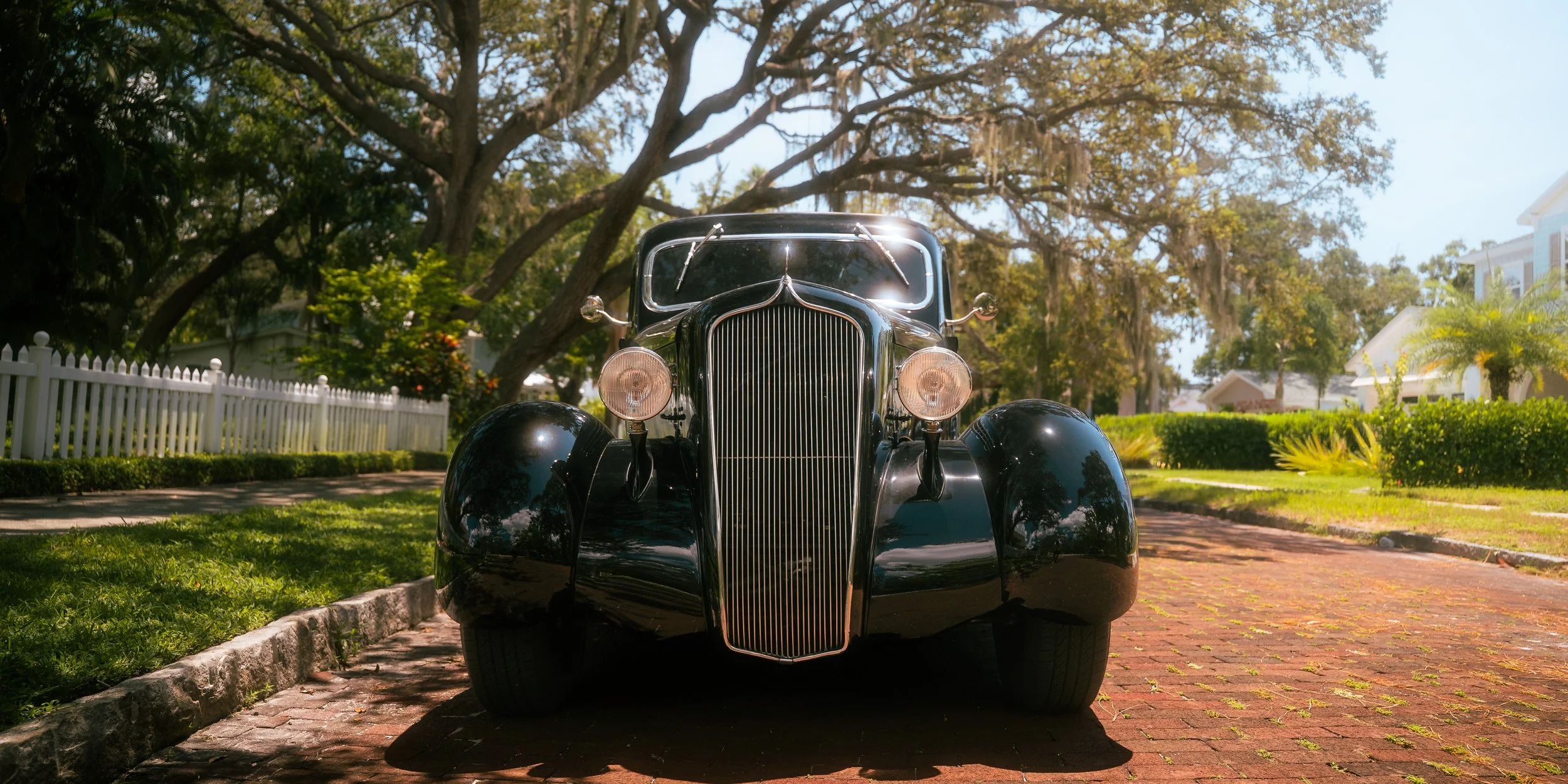 Front view of a vintage black car parked on a brick driveway surrounded by green grass and trees.