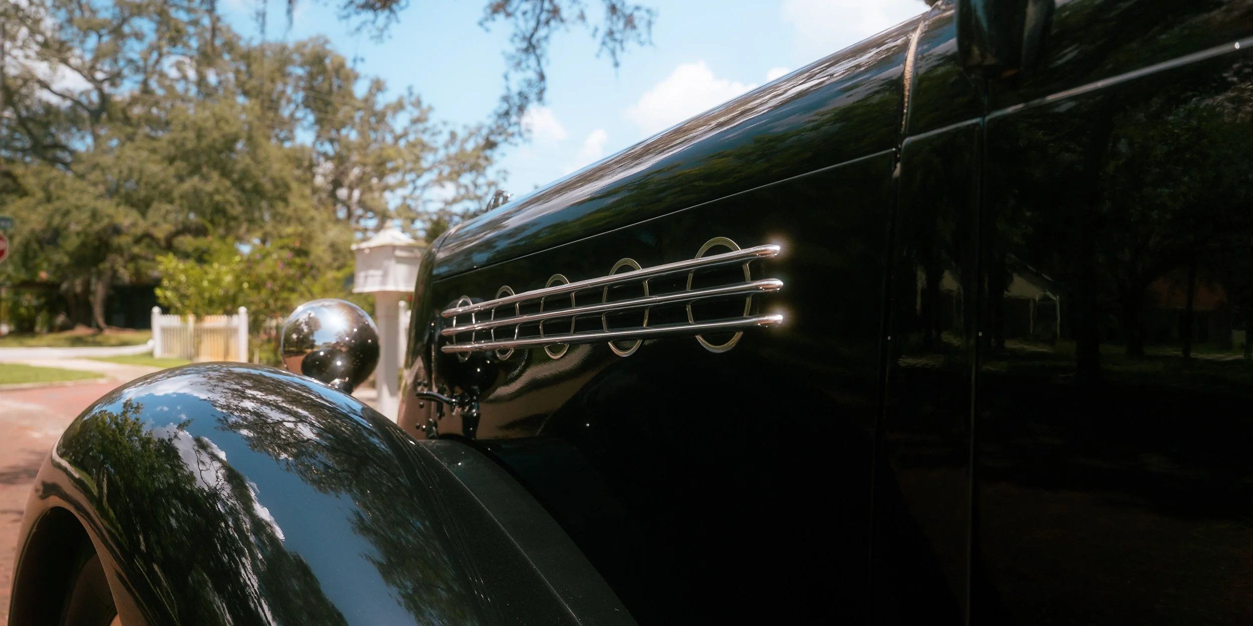 Close-up of a vintage black car with chrome accents and side vents, parked outdoors on a sunny day with trees and a house in the background.