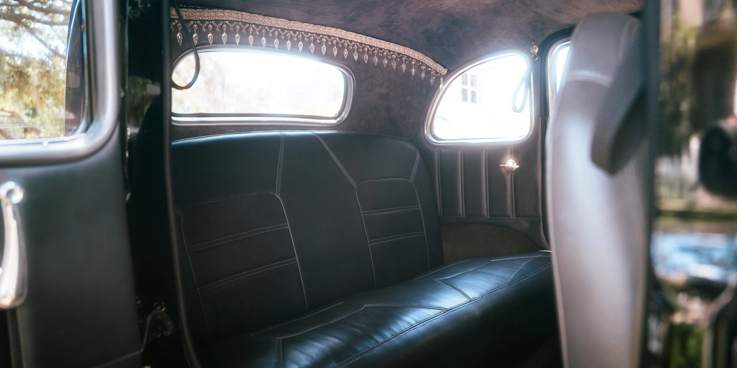 Interior of a vintage car with black leather seats, decorative trim along the window, and two small side windows showing outdoor trees.