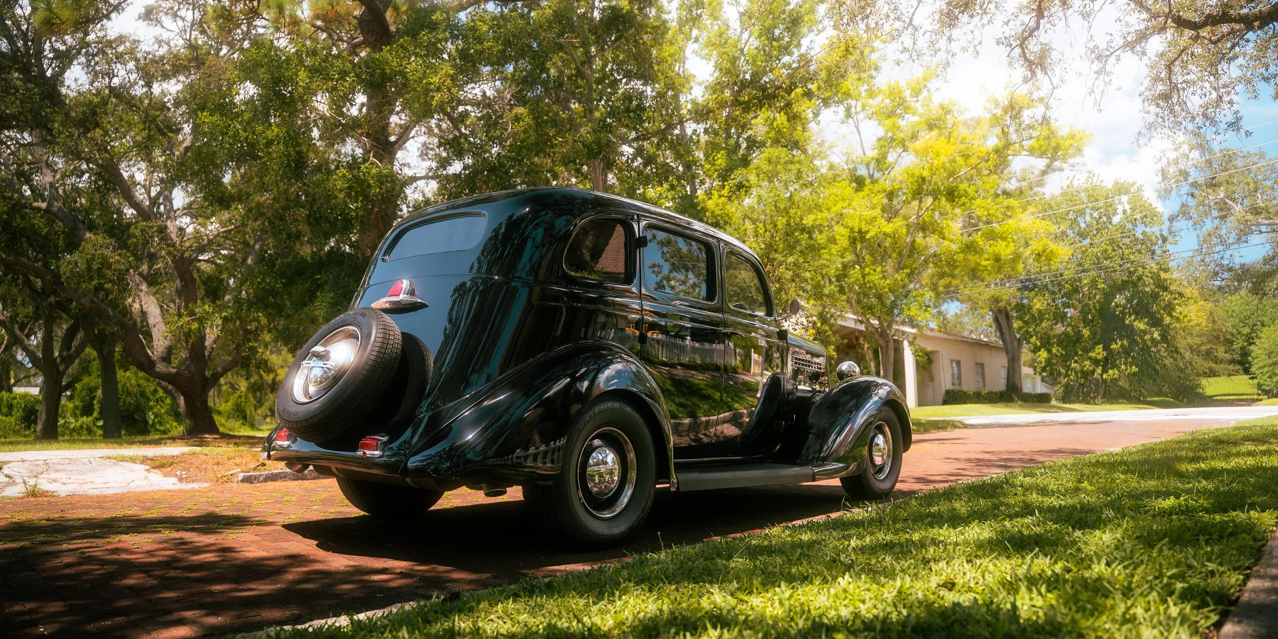 A vintage black car parked on a brick driveway in a suburban neighborhood with green trees and a white house in the background.